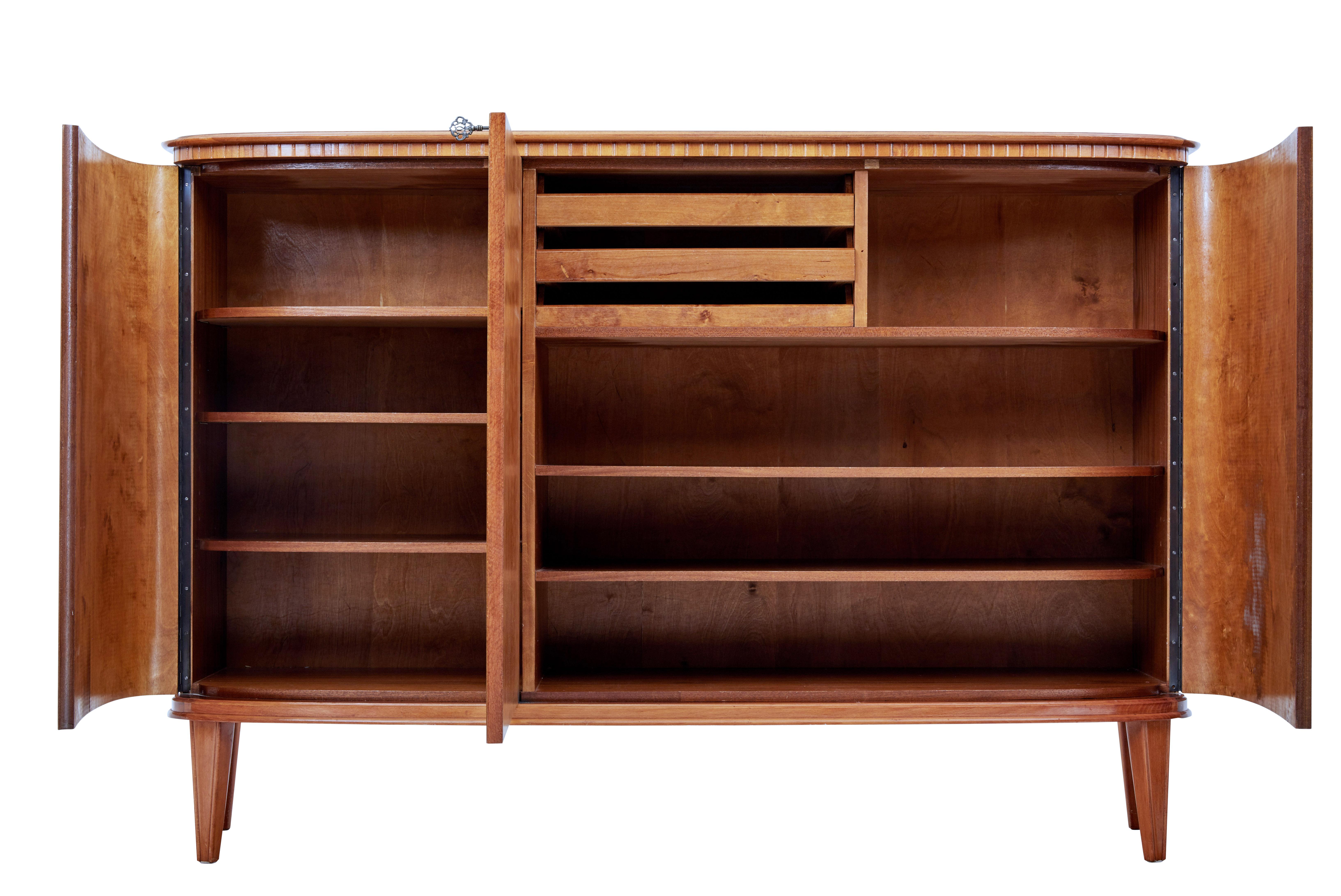 Striking 3-door sideboard, circa 1960.

Double door cupboard opens to reveal a partially fitted interior of 3-slide drawers and 3-shelves. Single door cupboard containing 3-shelves.

Matched flame mahogany used on the shaped doors with satinwood