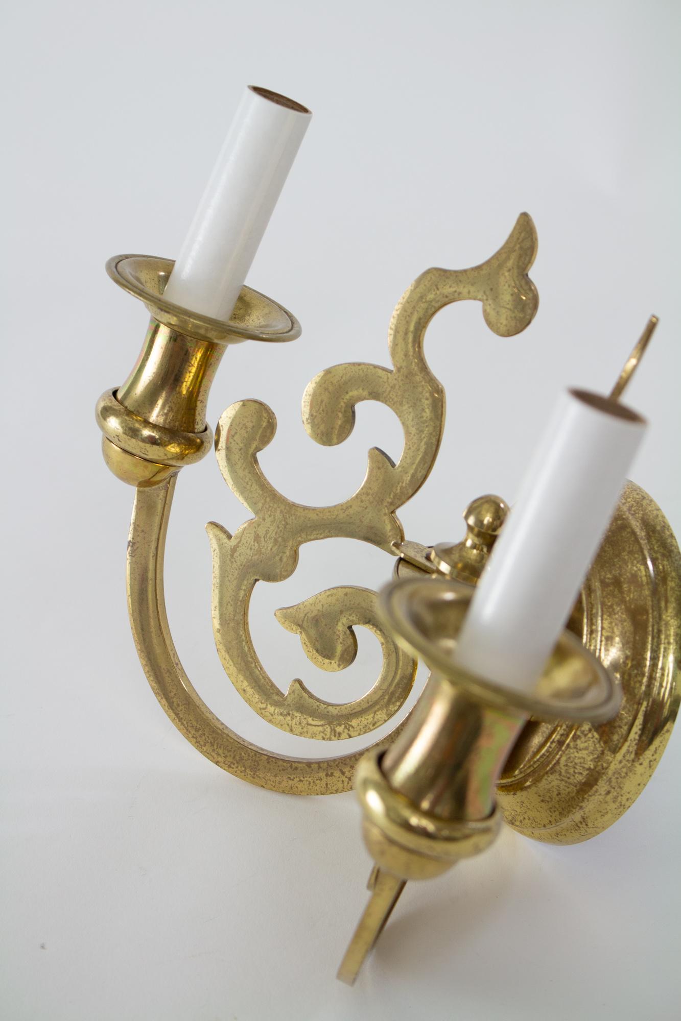 A pair of brass sconces, two arm, Hollywood regency style. Round Backplate. Two curved rectangular arms extend down and up from the center. Two flat brass winged ornaments extend above the arms. Central finial. Makers mark on on hanging hardware.