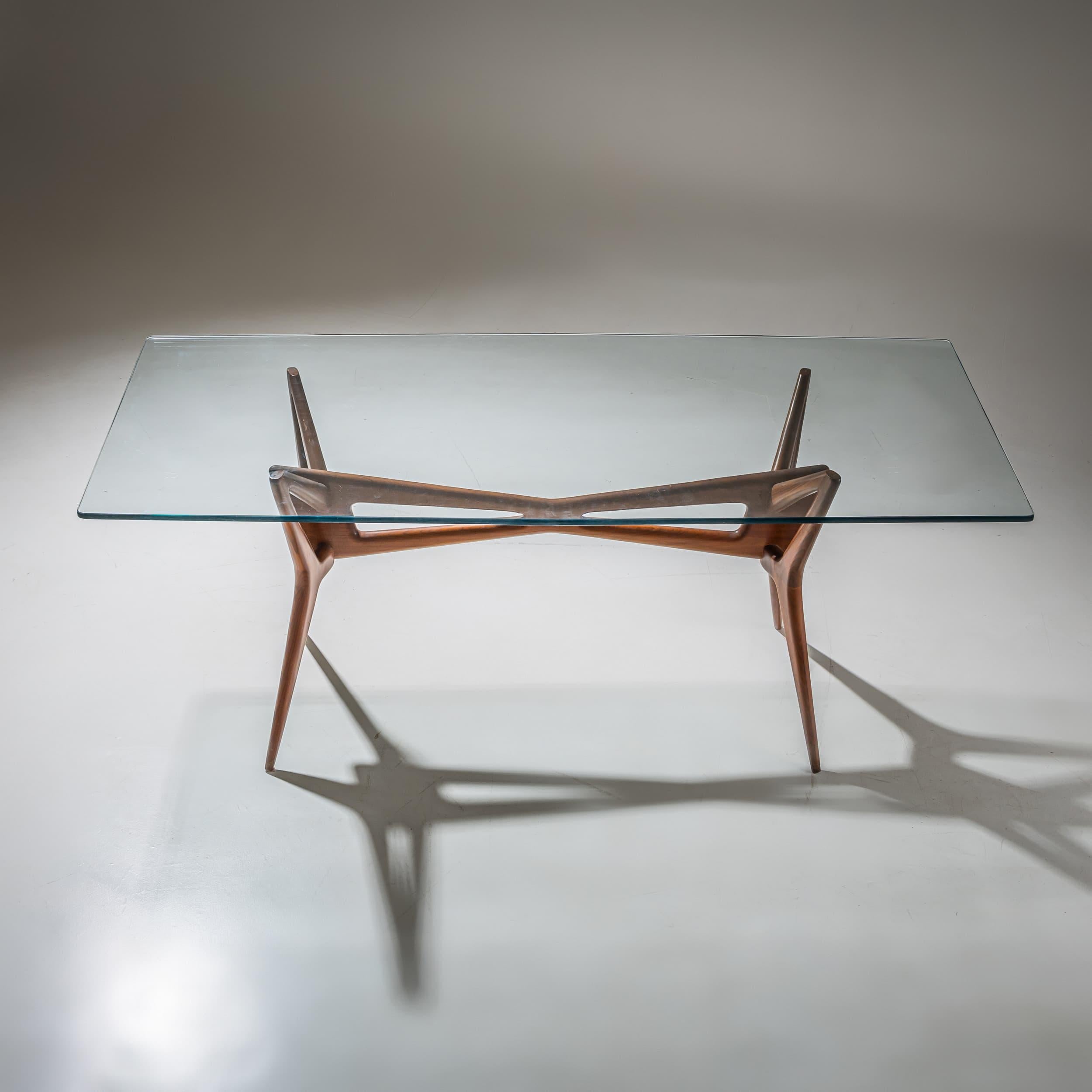 A refined dining table crafted in the 1950s. The sculptural wooden base features gracefully curved supports and sharply tapered, splayed legs, creating an elegant and airy presence. The rectangular glass tabletop rests delicately on four points,