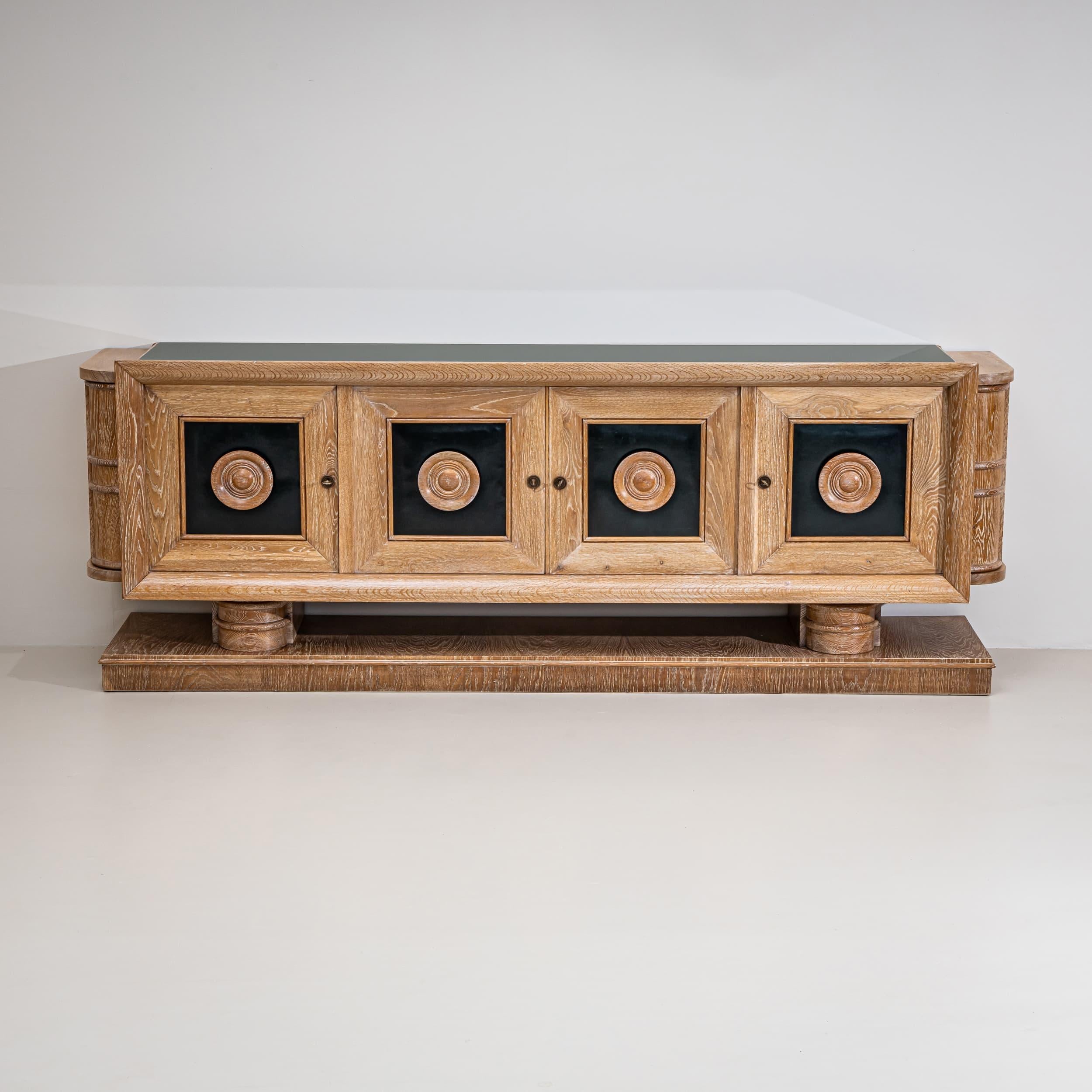 Grande credenza a quattro ante di Audoux-Minnet, realizzata in rovere sbiancato. I pannelli anteriori sono rivestiti in pelle verde scuro e il piano presenta una superficie in vetro verde scuro. Il pezzo poggia su una base rettangolare con due gambe