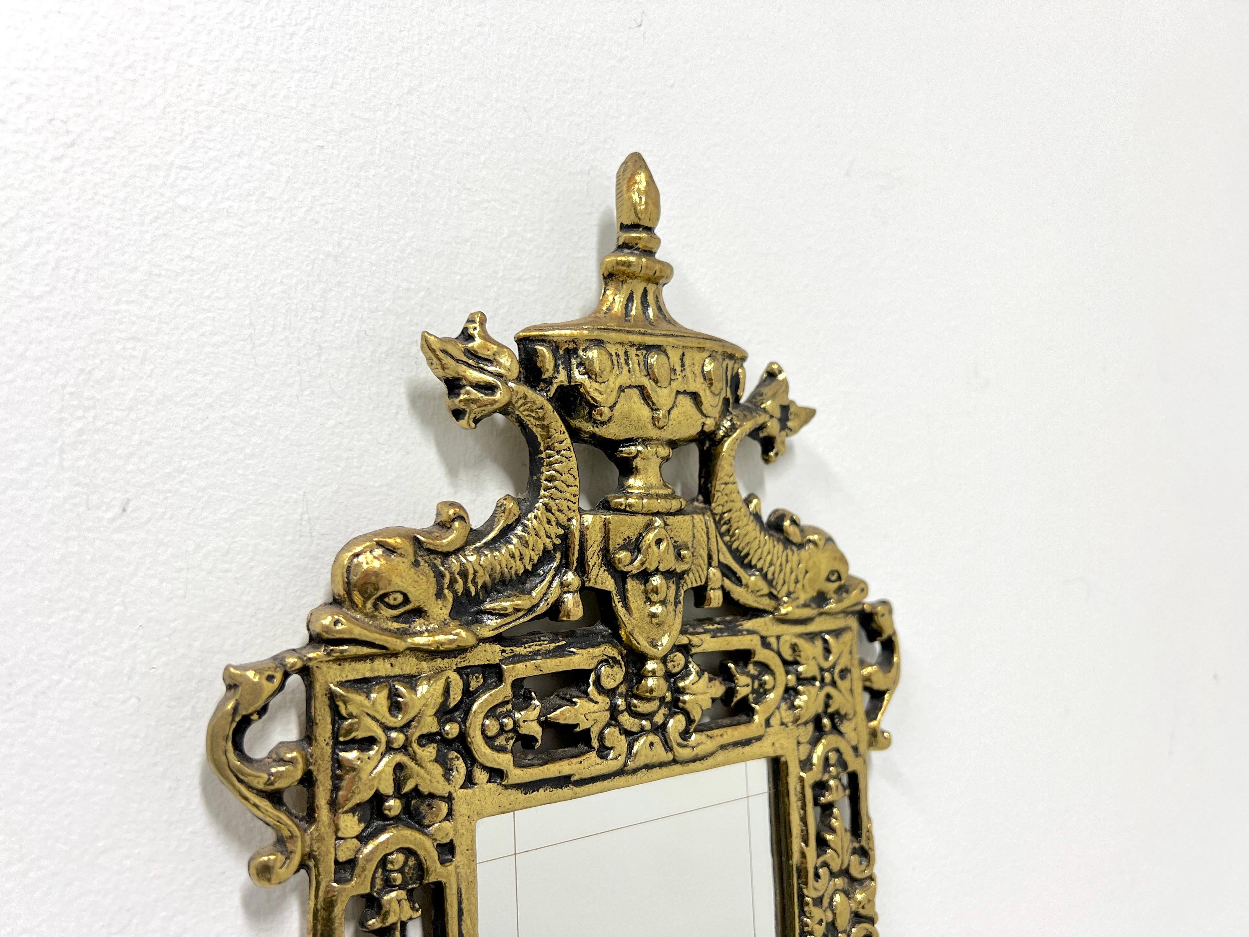 A French Provincial style mirrored candle wall sconce, unbranded. Solid brass decoratively sculpted with a center rectangular shaped mirror, urn shape with two mythical dolphins to the very top, decorative base, and two curved arms to support