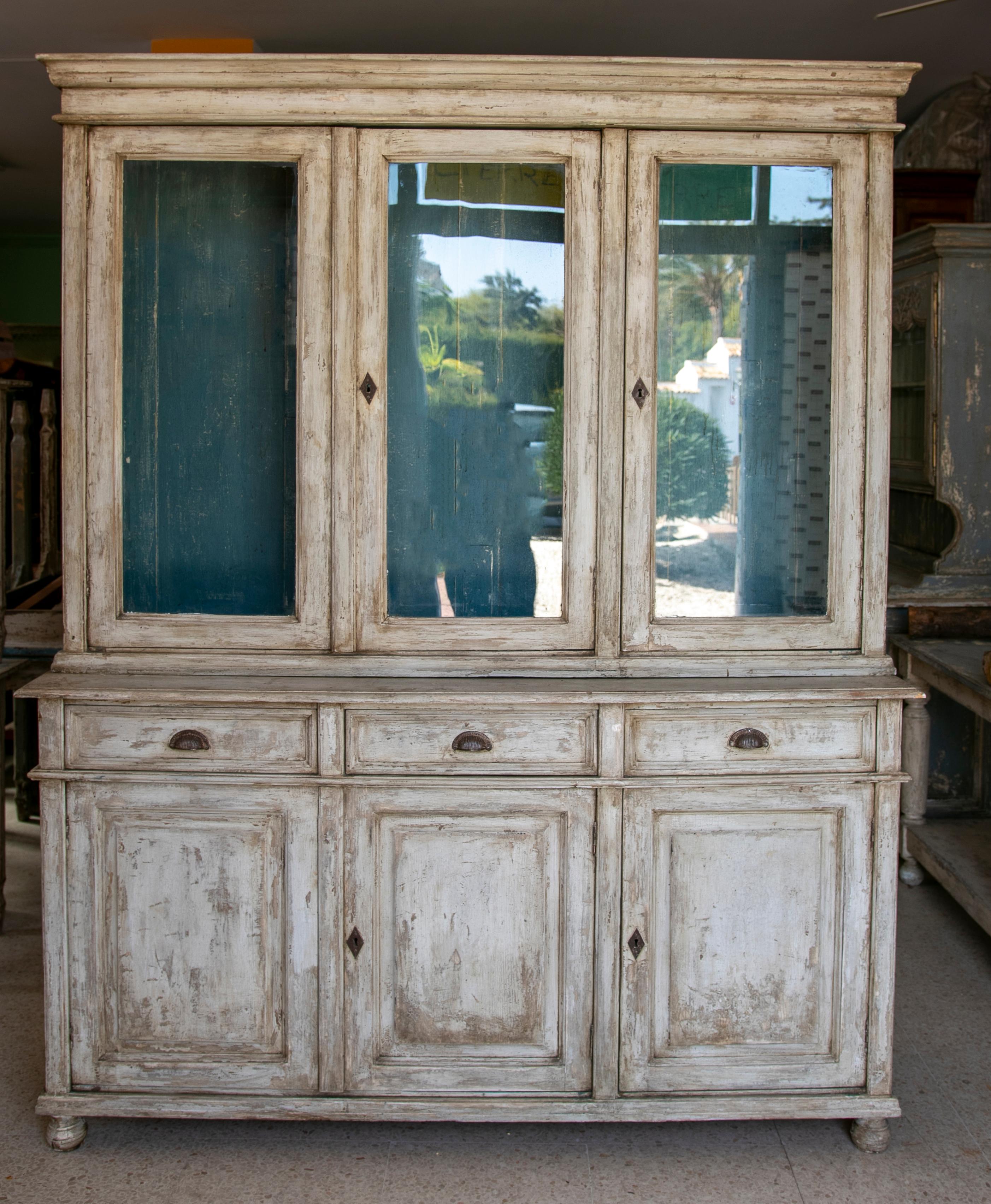 Rustic 1950s Spanish three glass panel door white painted bookcase cabinet with drawers and doors. The paint has worn off giving it a distressed weathered finish.
    