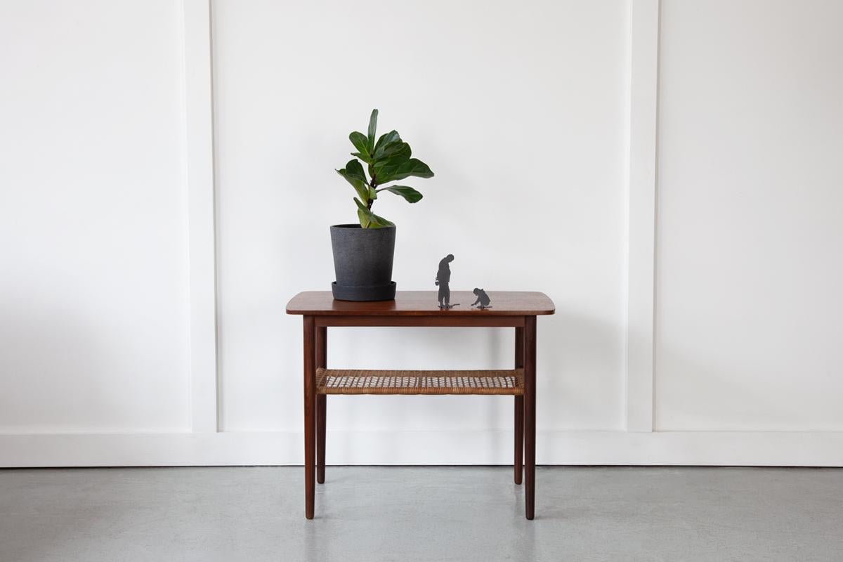 A sweet, Danish side table in teak with long, elegantly tapering legs and a useful magazine shelf made of woven cane. Perfect for use as a bedside table or living room side table. 
