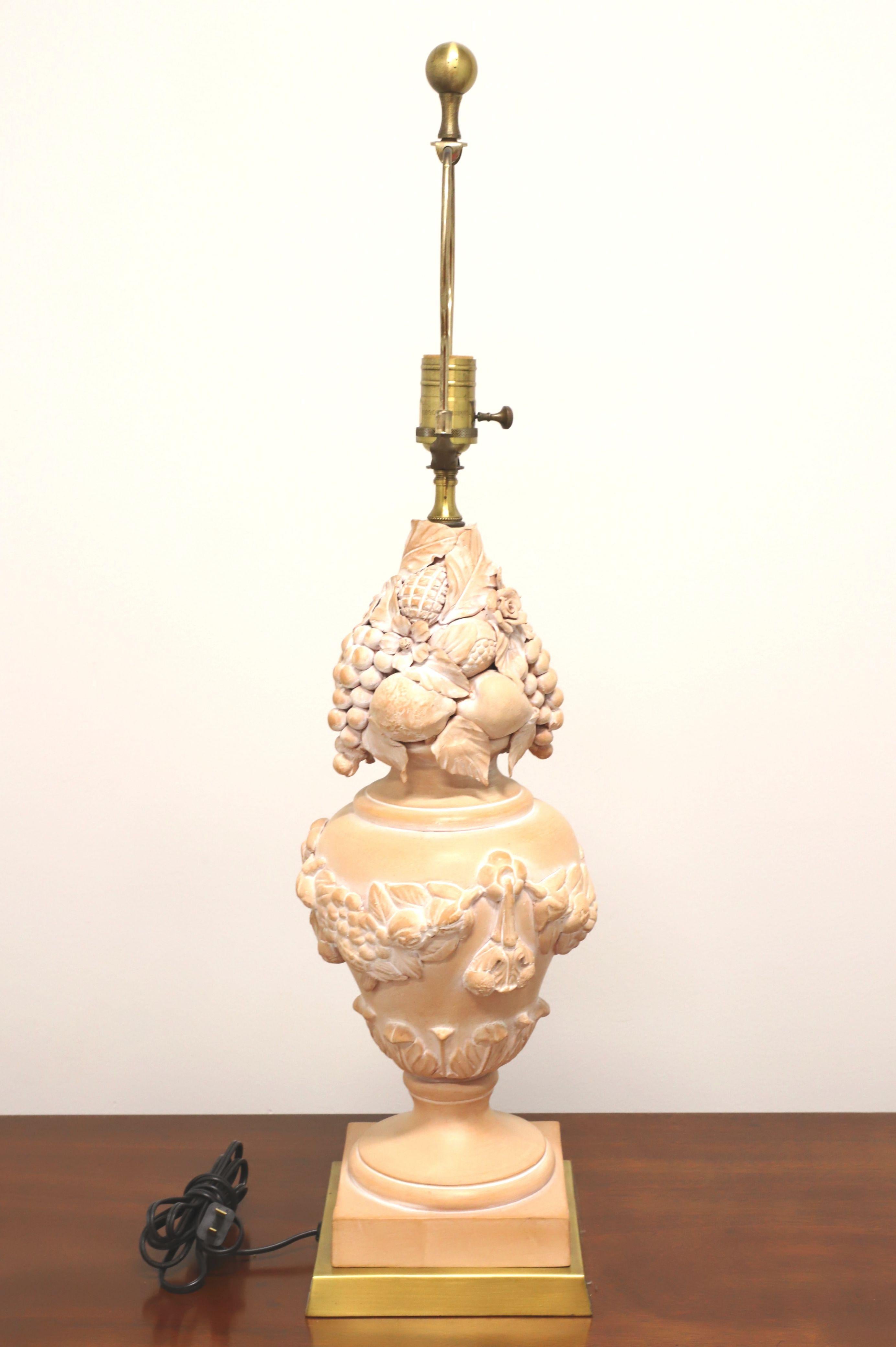 An Italian Tuscan style table lamp, unbranded. Made of terra cotta in an urn shape adorned with a fruit & floral motif, salmon & white in color, and a brass base. Has a removable brass harp and brass ball finial. Single standard bulb socket and