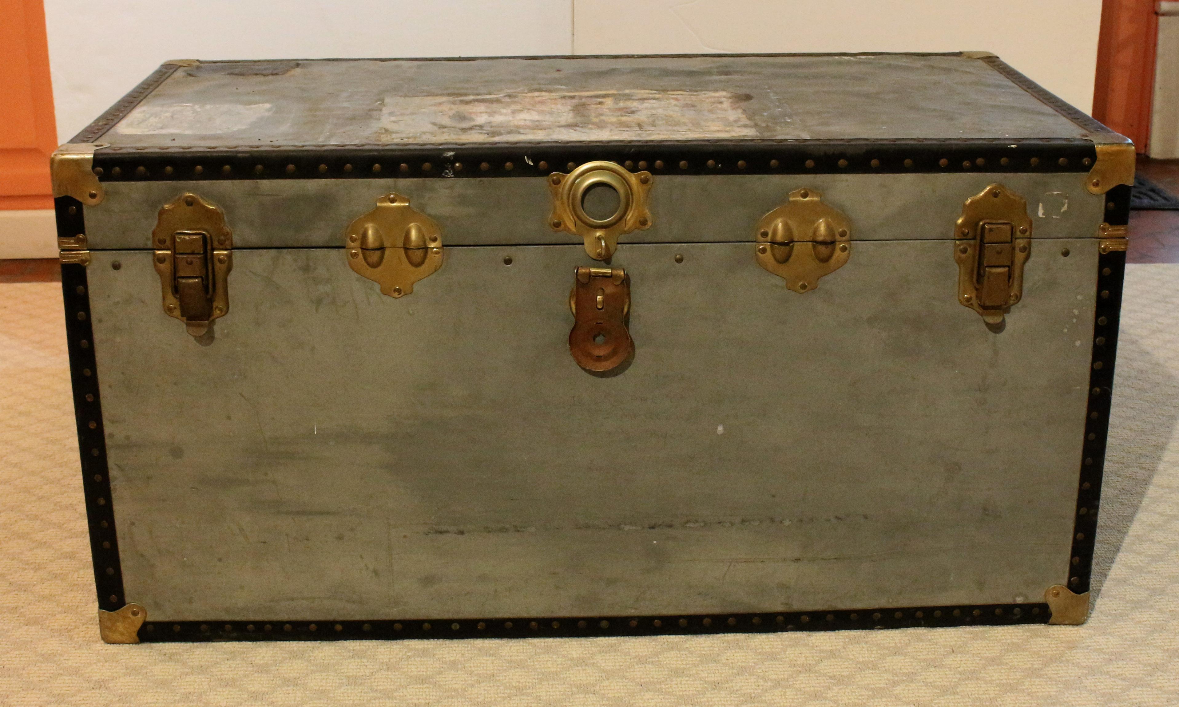 A zinc clad traveling trunk, Mid-20th Century. Wide black metal bindings, brass plated studs & fittings. One handle as is. Top as is. Measures: 39 3/4