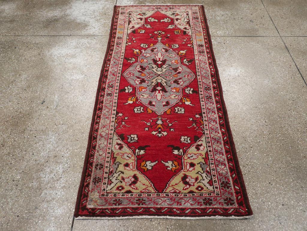Ein Wollflor mit symmetrischen Knoten auf einer Baumwollunterlage. Das rote Feld wird von einem länglichen Kartuschenmedaillon beherrscht, das in der Mitte ein Paar addierte Palmetten zeigt, an deren Rändern kleine Knospen wachsen. Die strohfarbenen
