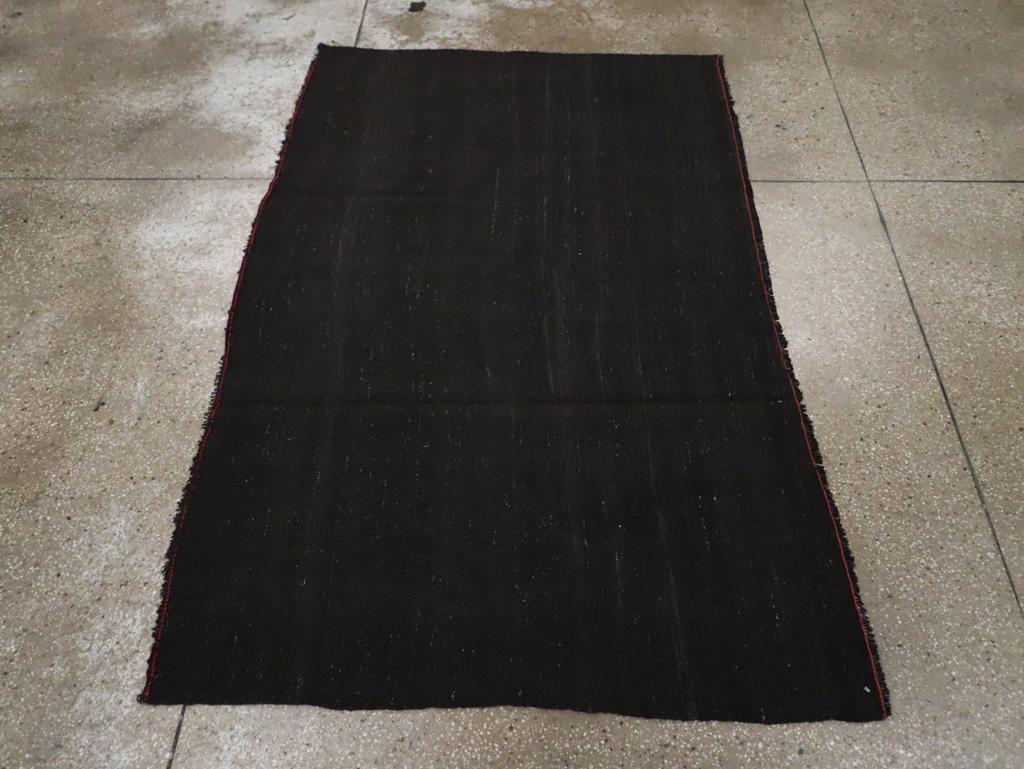 Esta alfombra Kilim turca de tejido plano de mediados del siglo XX, que mide 1,8 m x 1,8 m, presenta un diseño minimalista centrado en un campo negro profundo y entintado. La superficie monocromática se enriquece sutilmente con variaciones tonales