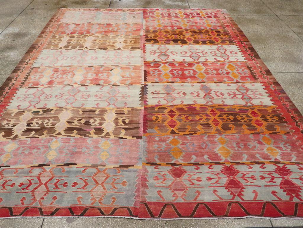 Esta alfombra Kilim turca de tejido plano de mediados del siglo XX, que mide 3 m x 4 m, muestra un diseño atrevido y expresivo, rico en motivos tribales tradicionales. La alfombra está dividida en dos paneles verticales, cada uno lleno de bandas