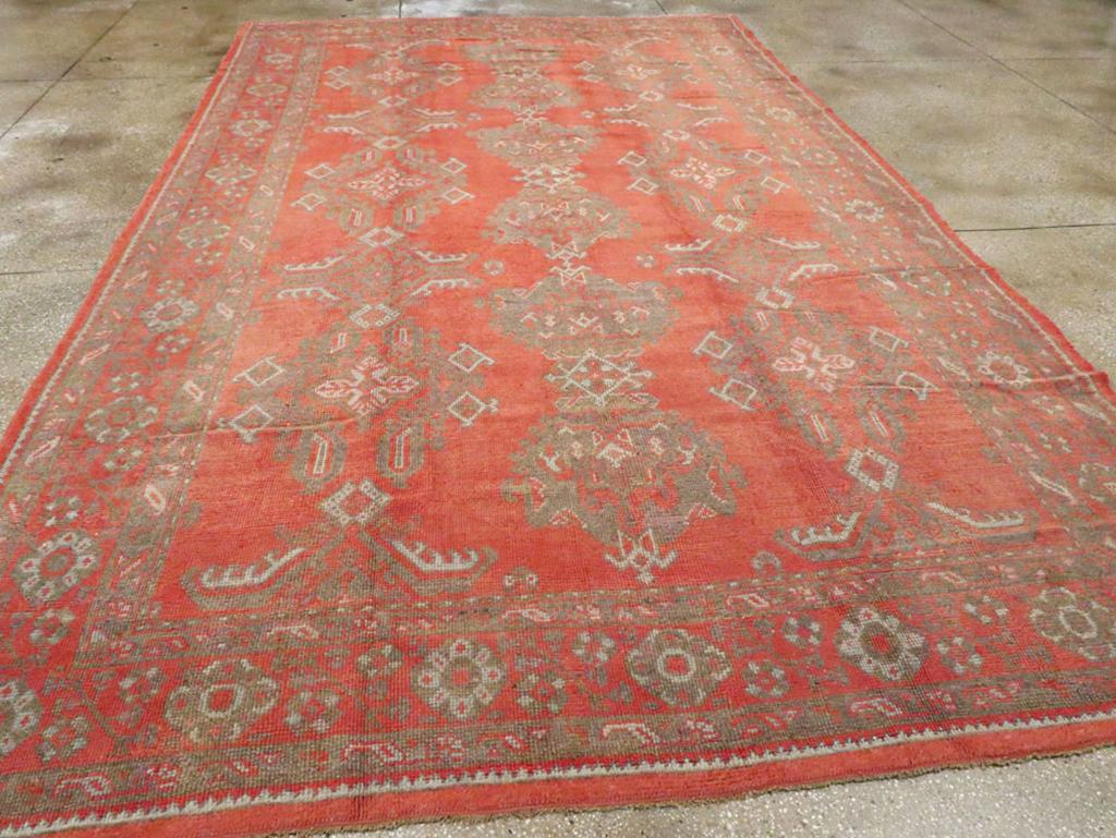 A vintage Turkish Oushak room size carpet. The light red field of this antique Oushak displays a central vertical pole medallion of connected Harshang palmettes set between columns of lozenges, crosses and bent stiff leaves (Yaprak). This overall