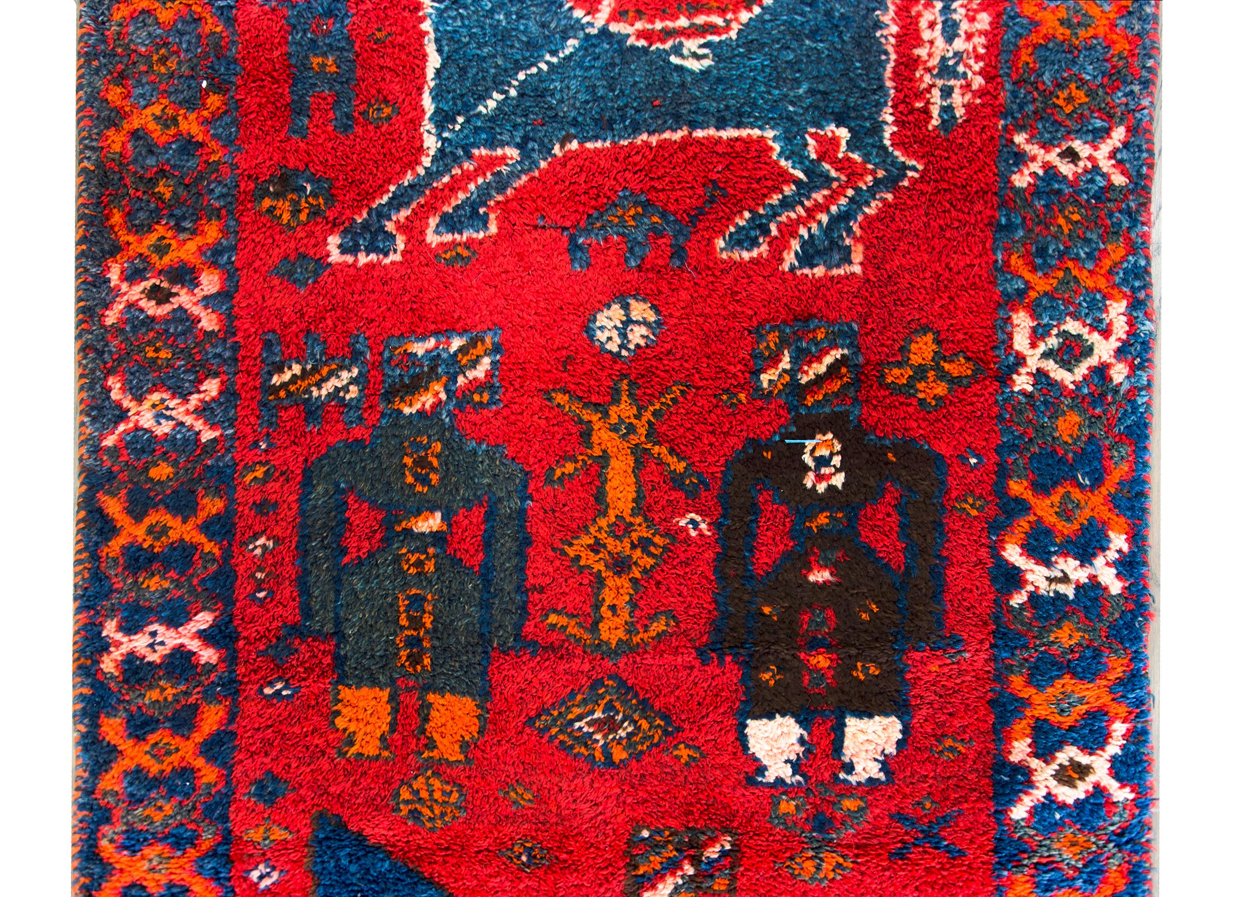 A bold mid-20th century Turkish Oushak rug with an unusual pattern containing standing figures and two figures on horseback all woven in brilliant indigos, turquoise, orange, and white set against a crimson background, and surrounded by a geometric