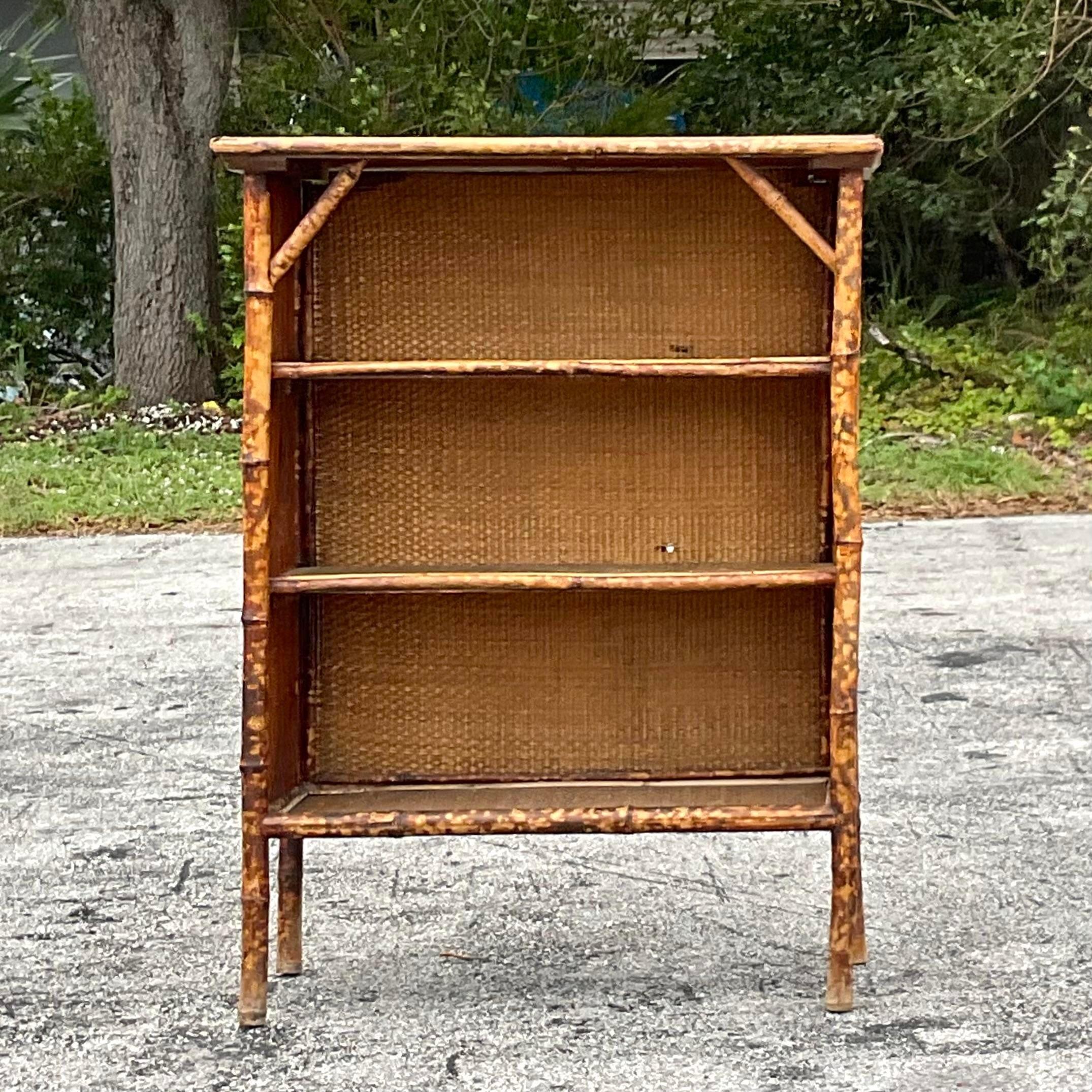 A fantastic vintage Boho bookshelf. A chic burnt bamboo frame with inset woven rattan panels. A hand painted lacquered floral top. Acquired from a Palm Beach estate.