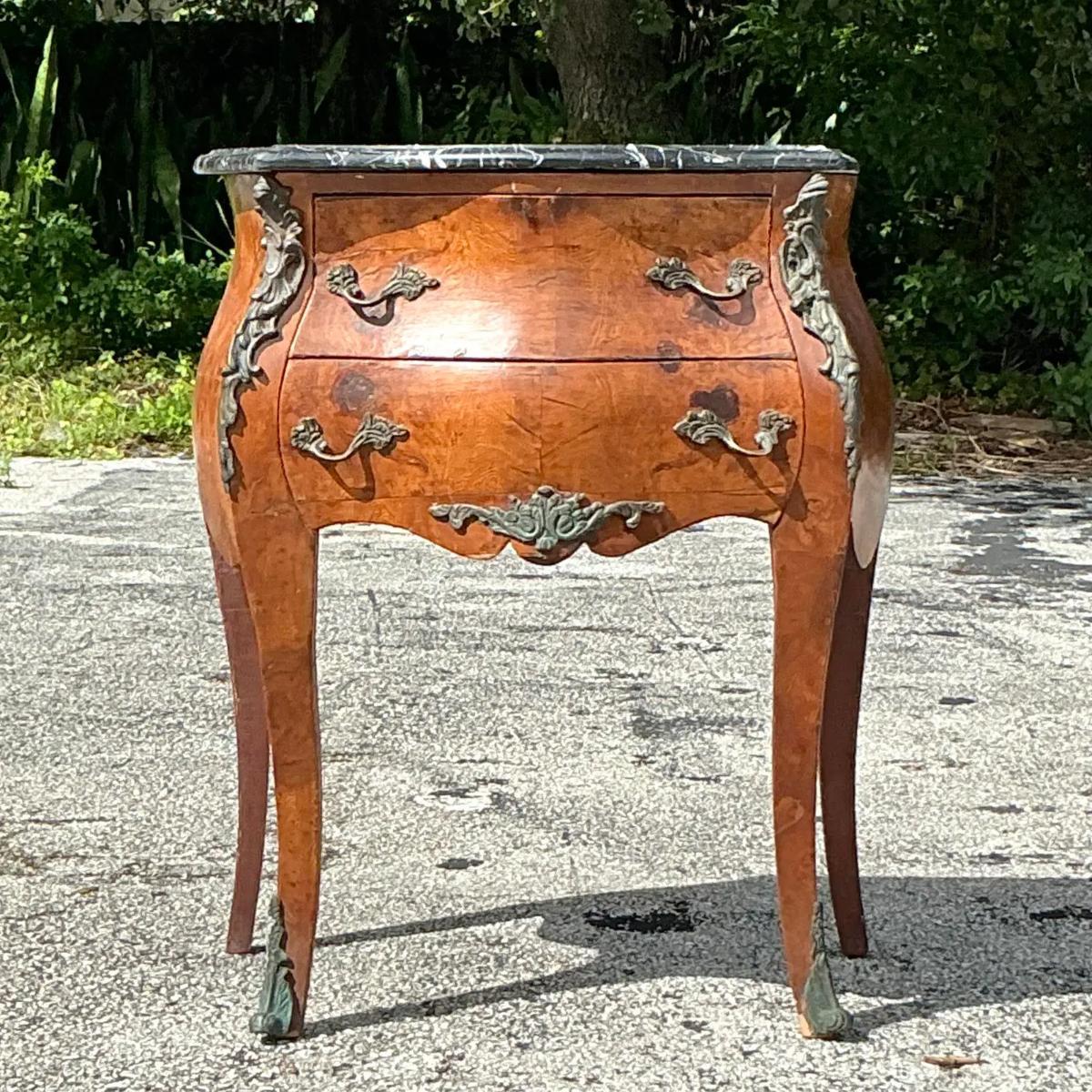 Cette commode en bois de ronce et bronze doré de style Vintage Regency exsude le luxe. Fabriqué avec un placage de loupe exquis et orné de touches ornementales en bronze doré, ce coffre est une déclaration d'élégance intemporelle. Ses riches