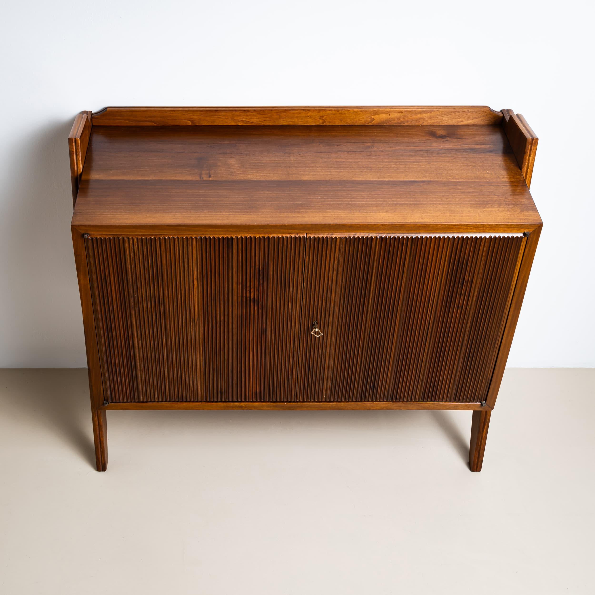 Elegant walnut sideboard, crafted in Italy in the 1950s. The distinctive fluted double doors and wedge-shaped side supports create a striking architectural profile.
Inside, a single shelf provides practical storage space. A low gallery rail along