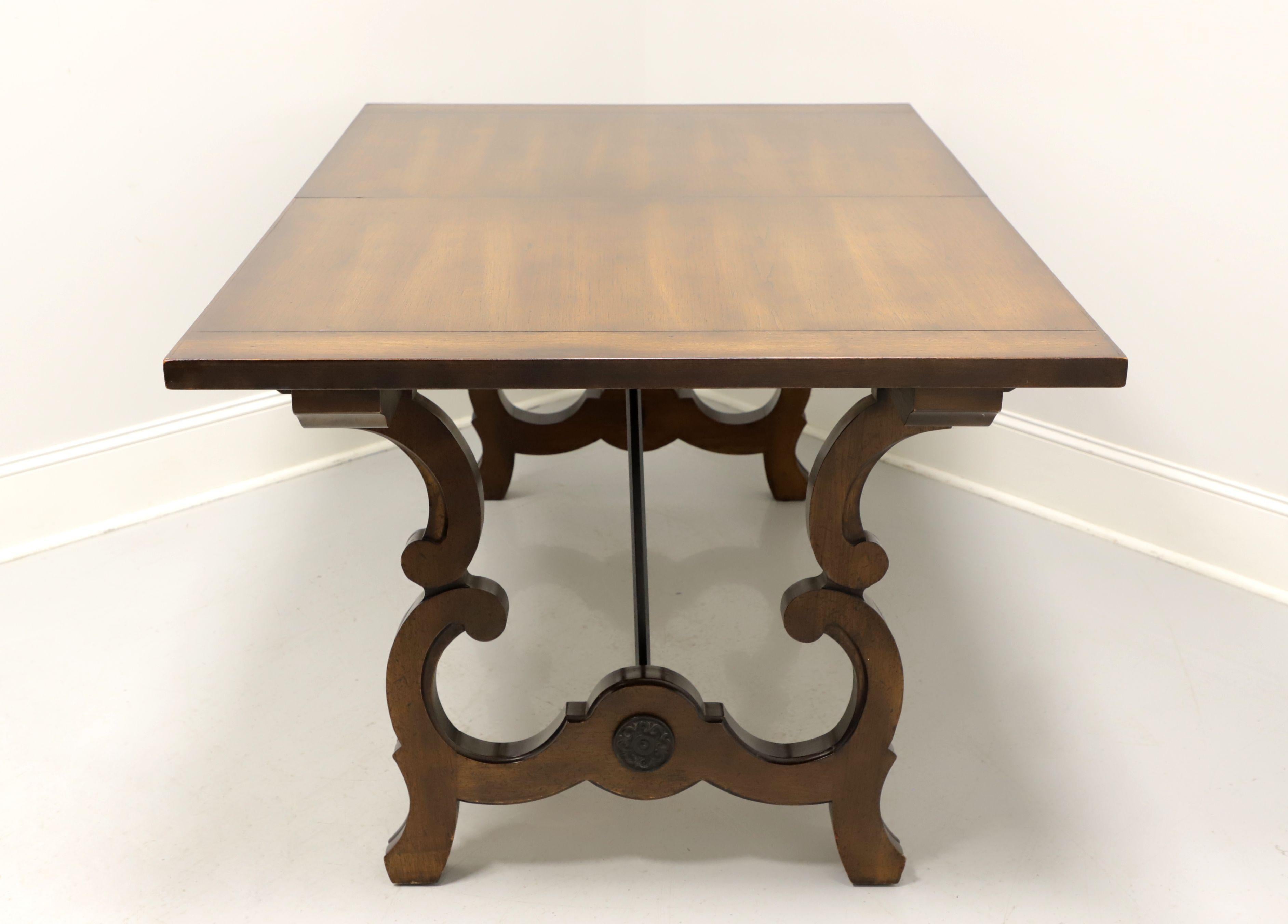 A Spanish Baroque style dining table, unbranded. Walnut with banded ends to top, decoratively carved trestle base legs with black metal rods support and decorative metal medallions to ends. Wood expansion sliders. Includes two extension leaves. Made