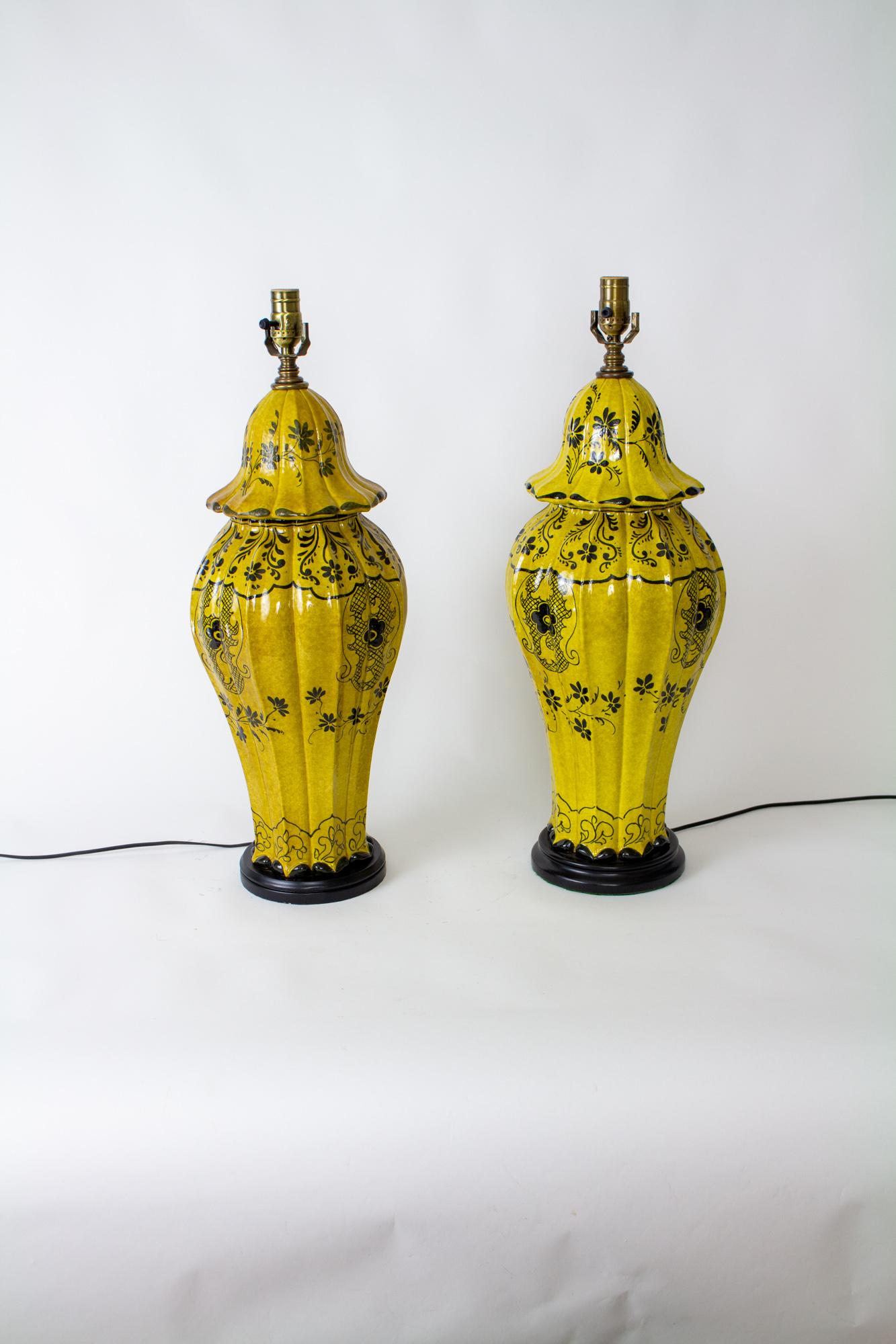 A large pair of ceramic table lamps. Vase shaped with lids, a ribbed shape. Mustard colored glaze with hand-drawn black accents and black wooden bases. Design is of flowers and leaves. Rewired with new sockets. The lamps are slightly varied in