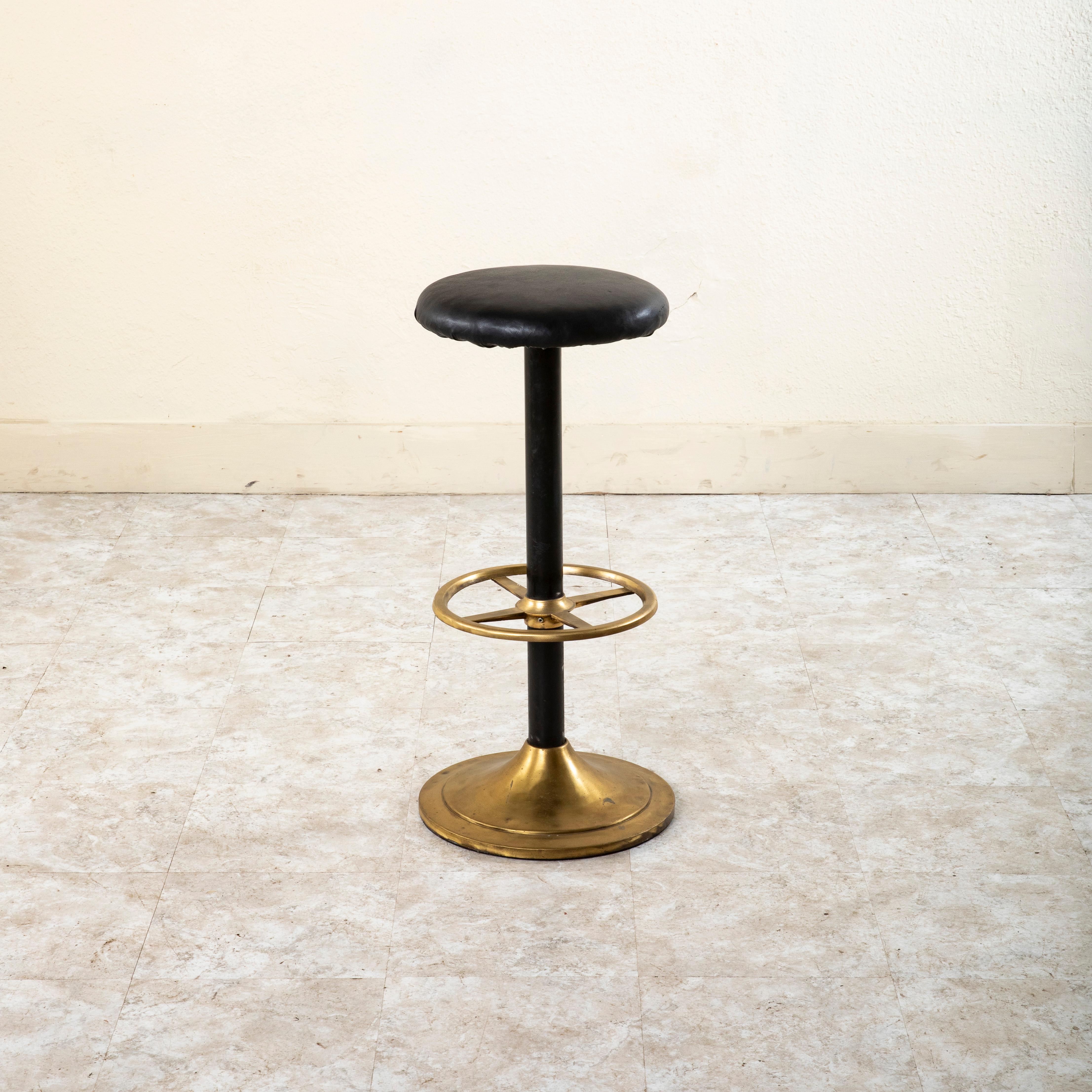 Questo sgabello da bar francese in ottone della metà del XX secolo presenta una base circolare in ottone con una colonna centrale in metallo nero. Un poggiapiedi in ottone è unito alla colonna centrale con quattro supporti. La seduta dello sgabello