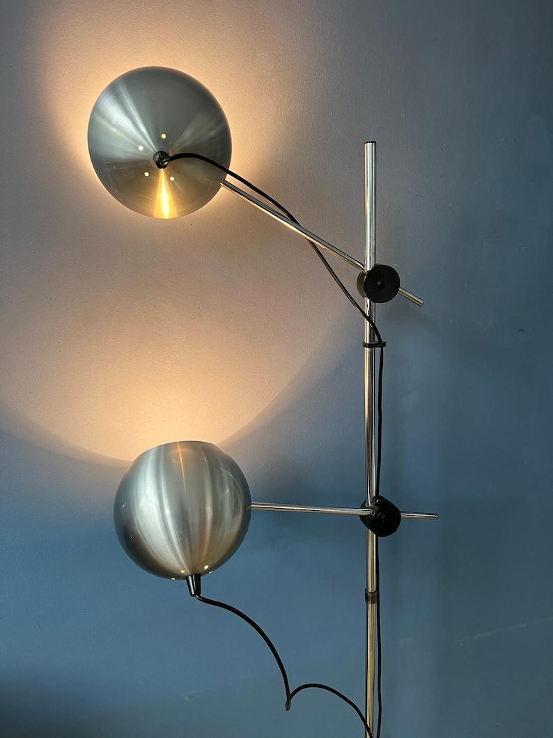 Lampada da terra a forma di bulbo oculare in alluminio con doppi bracci oscillanti. I bracci girevoli che trasportano i paralumi possono muoversi su e giù per la base ed essere posizionati in diverse angolazioni. La lampada richiede due lampadine