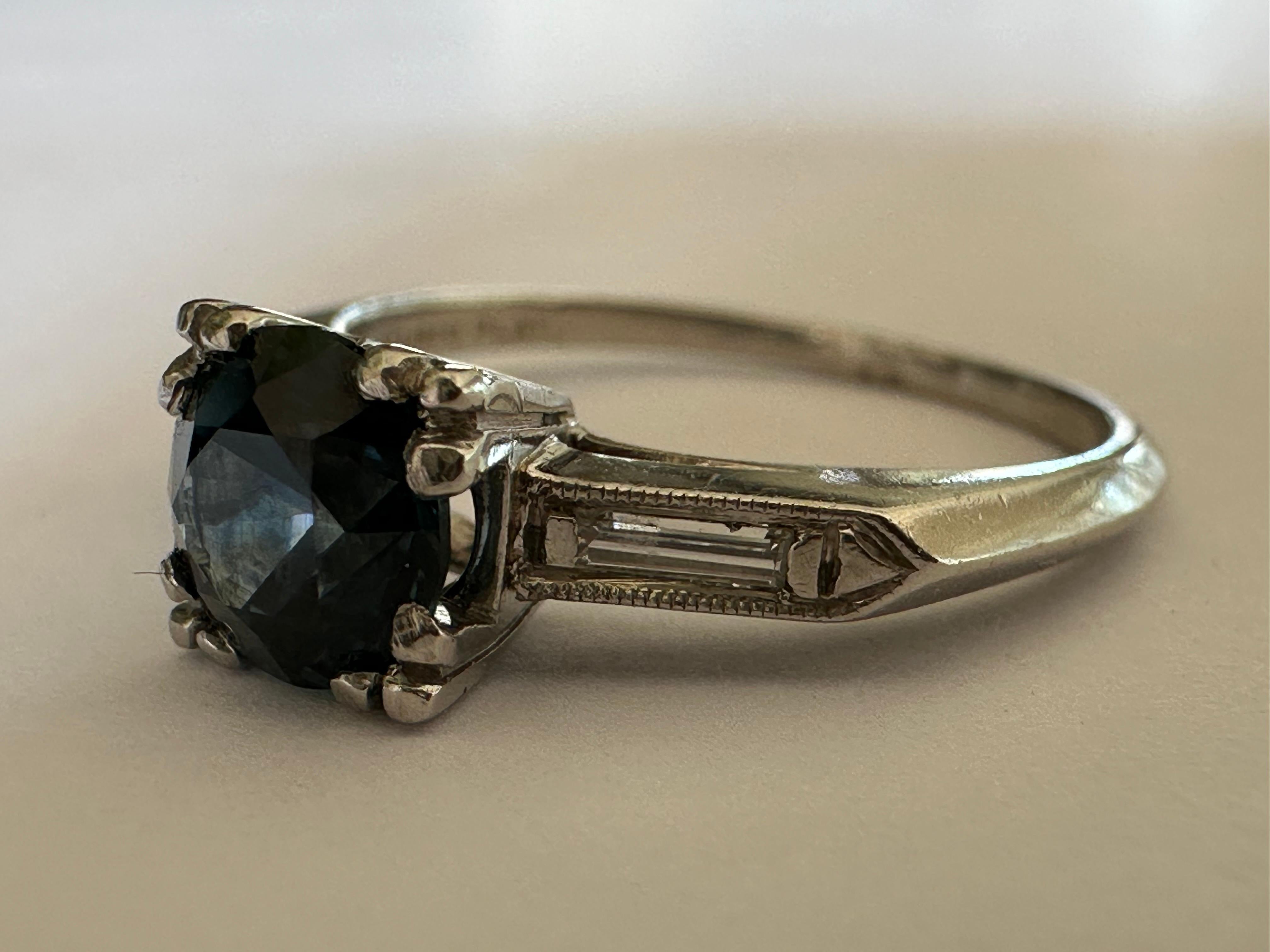 A rare 1.08-carat natural deep blue Montana sapphire measuring 6.05 mm centers this mid-century ring crafted in the 1950s flanked by two baguette diamonds totaling approximately 0.10 carat and bordered by fine millegrain edging. Set in platinum. 

