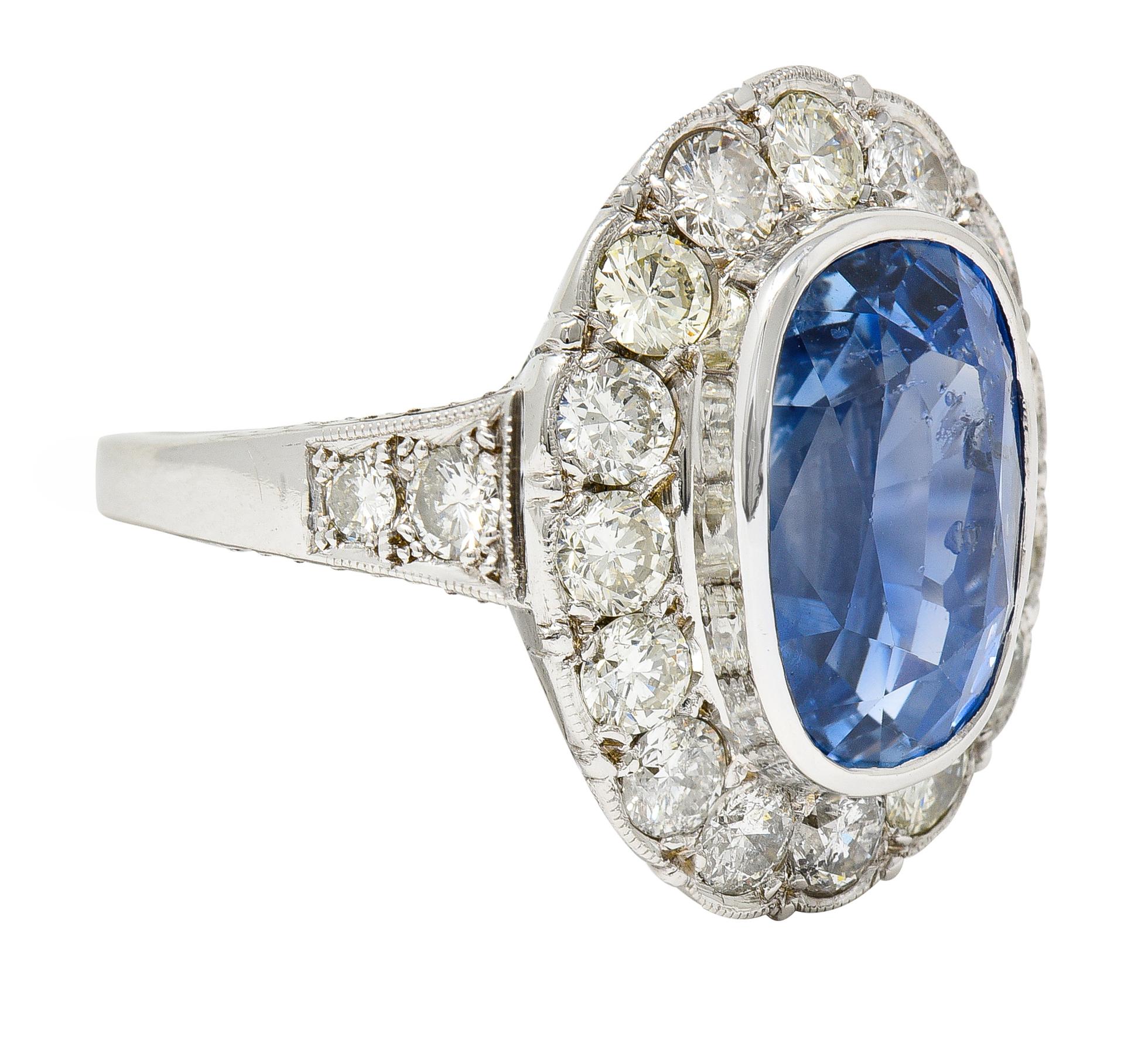 Centering an elongated cushion cut sapphire weighing 8.50 carats - transparent medium blue. Natural Sri Lankan in origin with no indications of heat treatment - bezel set. Featuring a scalloped halo surround of bead set transitional cut diamonds.