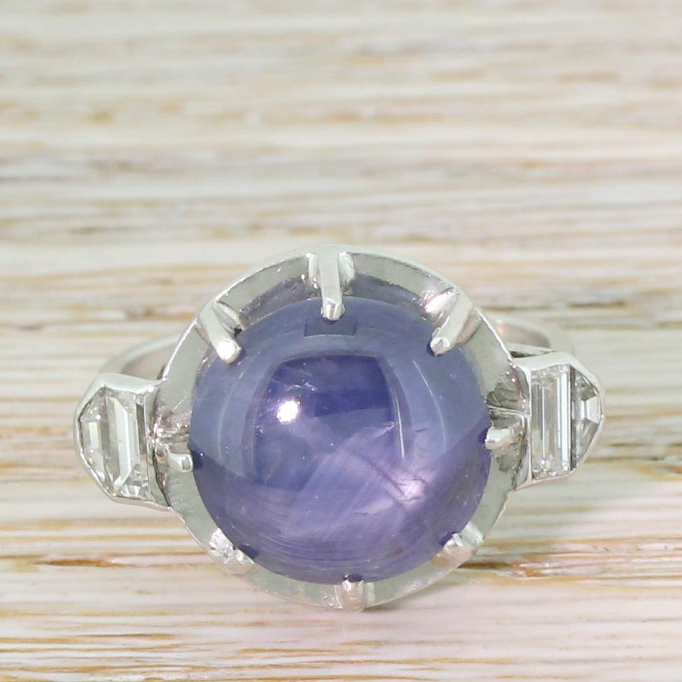 An impressive star sapphire takes centre stage in this beautifully unique ring. The bright, denim blue sapphire displays a clear asterism and is showcased in an eight claw open collet above a smooth – almost mirrored – backplate. Each shoulder is