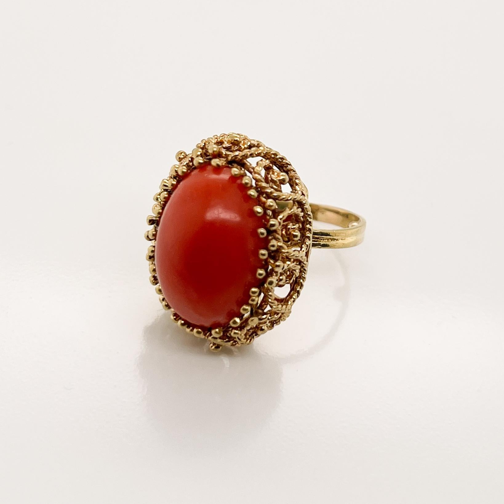 A very fine 14 karat gold and coral cocktail ring.

With a large oval coral cabochon prong-set in 14k gold.  

The bridge and base of the setting are made of twisted wire shaped to form a filigree pattern and are supported on a fused wire shank.

A