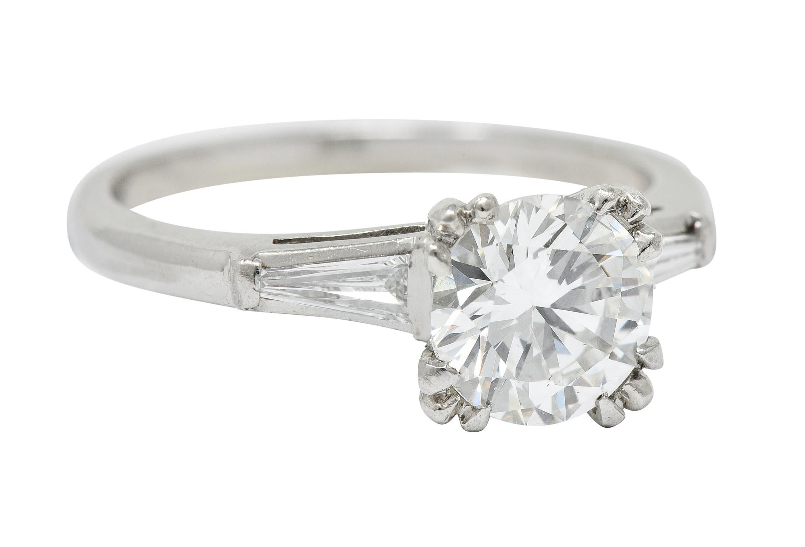 Centering a round brilliant cut diamond weighing 1.18 carat - F color with SI2 clarity

Set by tri-split talon prongs in a stylized square form head

Flanked by bar set tapered baguette cut diamonds weighing in total approximately 0.22 carat -