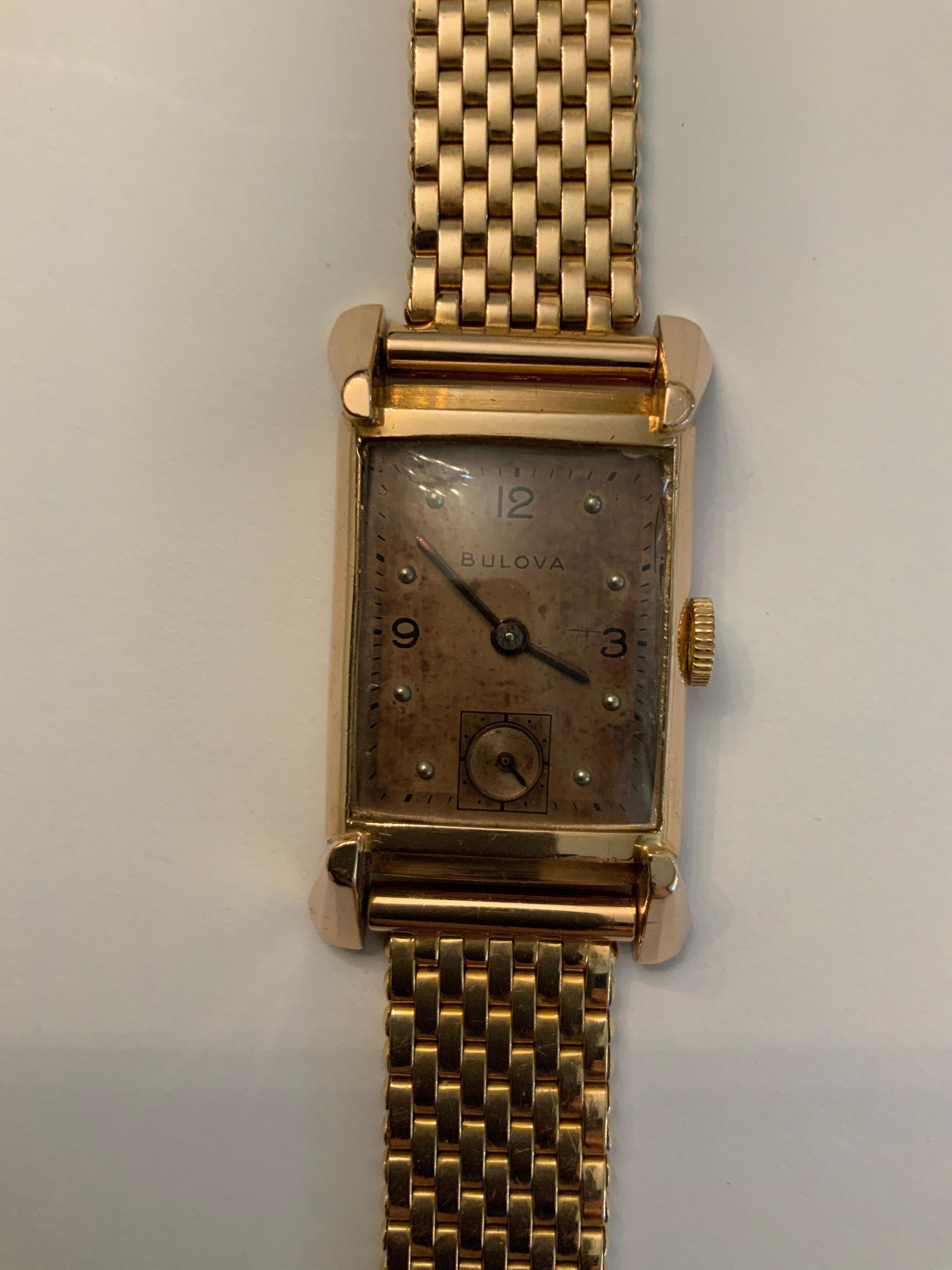 Se trata de un reloj Bulova de mediados de siglo con caja de tanque ROSA de 14 quilates. Representa un reloj de pulsera de oro rosa con una caja rectangular cubierta por un cristal. Tiene una pulsera estilo Jubileo de oro de 14 quilates. En la