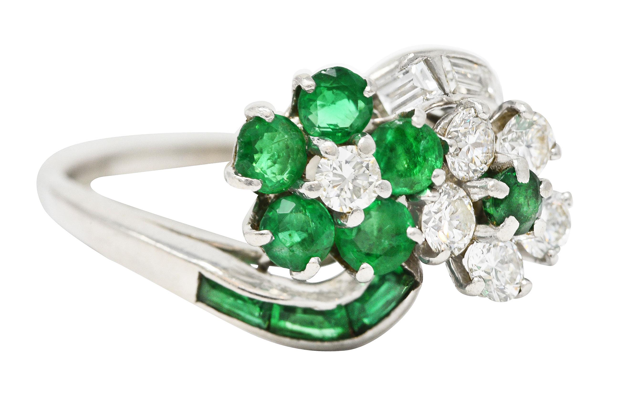 Designed as a bypass style ring terminating with two floral motif clusters of prong set emeralds and diamonds. With additional baguette cut emeralds and diamonds channel set in shoulders. Emeralds weigh approximately 0.73 carat total - transparent