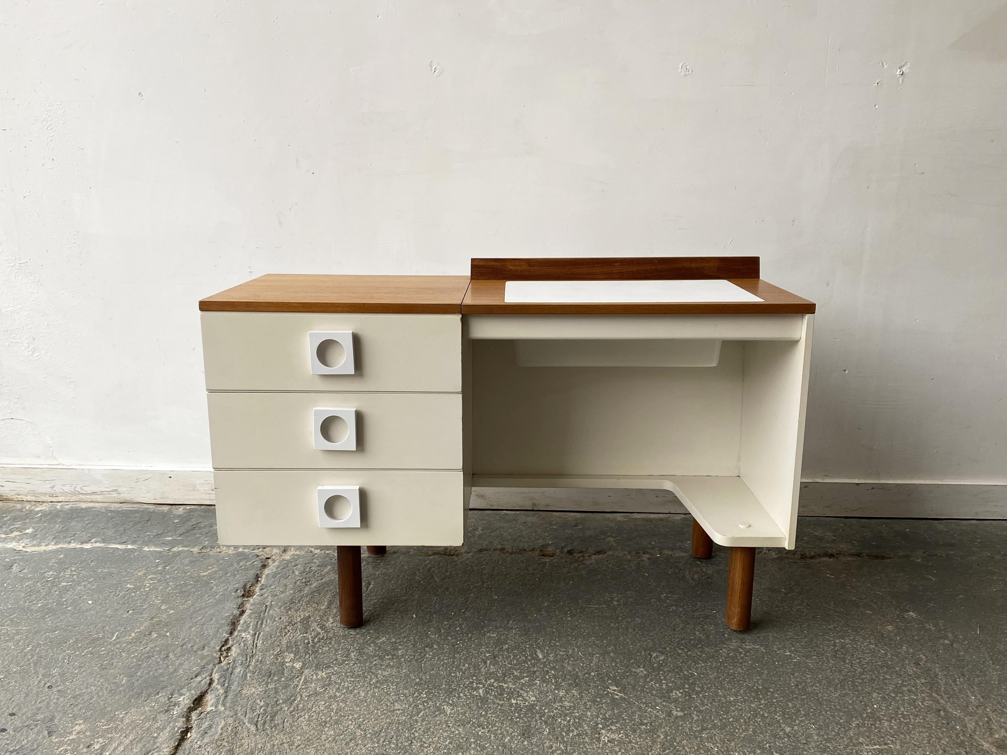 Una inusual y rara combinación de escritorio y tocador del fabricante inglés Uniflex. Con la solapa bajada  es una mesa de trabajo, levanta la solapa para descubrir un espejo y se convierte en un tocador. Contraste de melamina blanca y teca de color