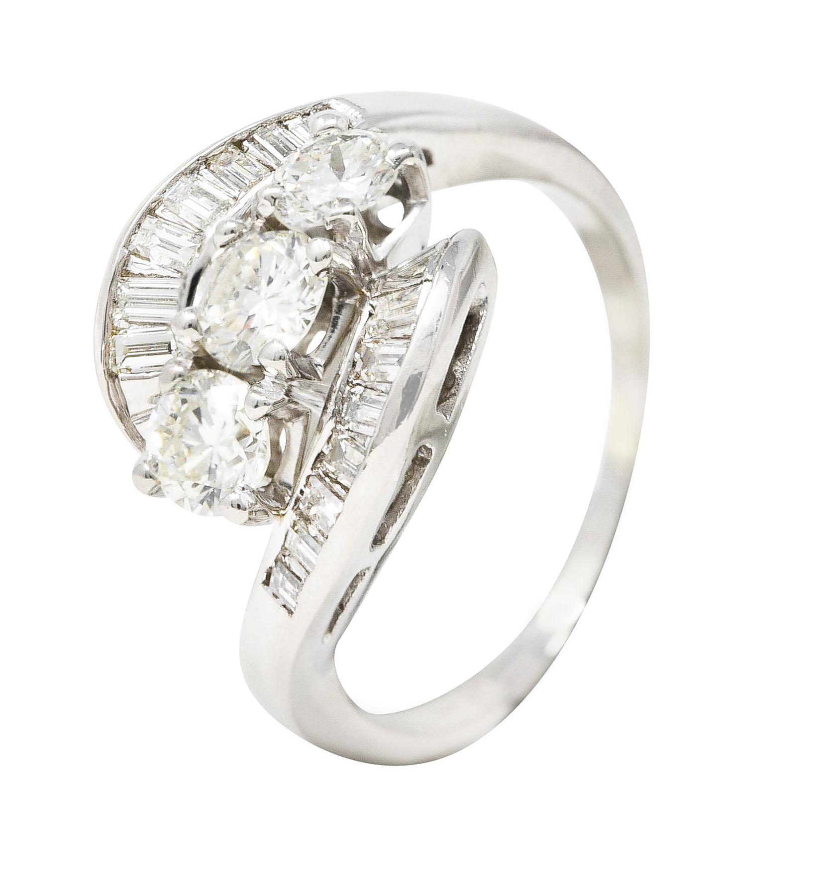 Centering three round brilliant cut diamonds prong set East to West. Weighing approximately 1.11 carats total - I/J in color with SI clarity. Flanked North to South by swirling bypass style shoulders. Channel set with baguette cut diamonds