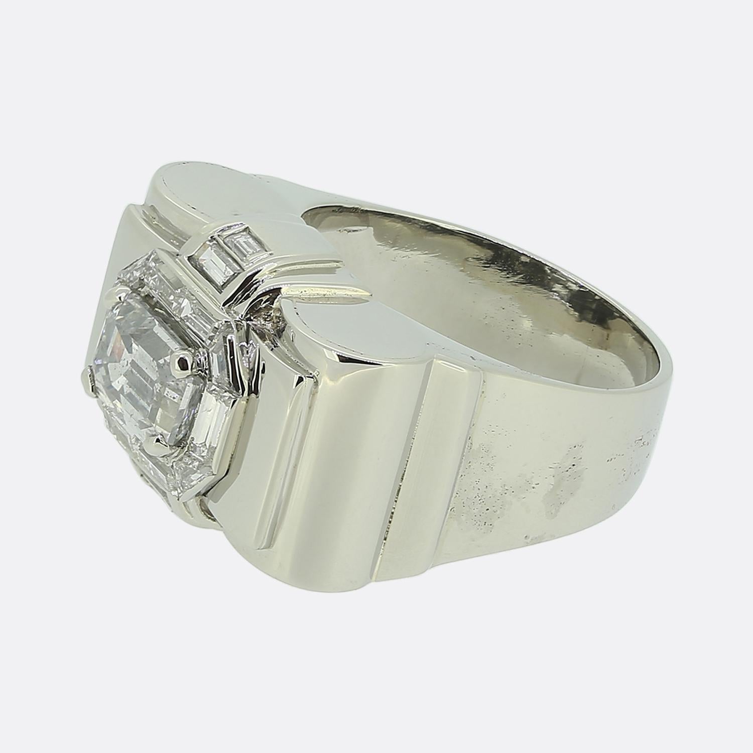 Aquí tenemos un anillo de platino de mediados del siglo XX. Esta pieza vintage presenta en el centro un diamante blanco talla esmeralda. Esta piedra principal está ligeramente elevada en un engaste de cuatro garras, enmarcada por una serie de