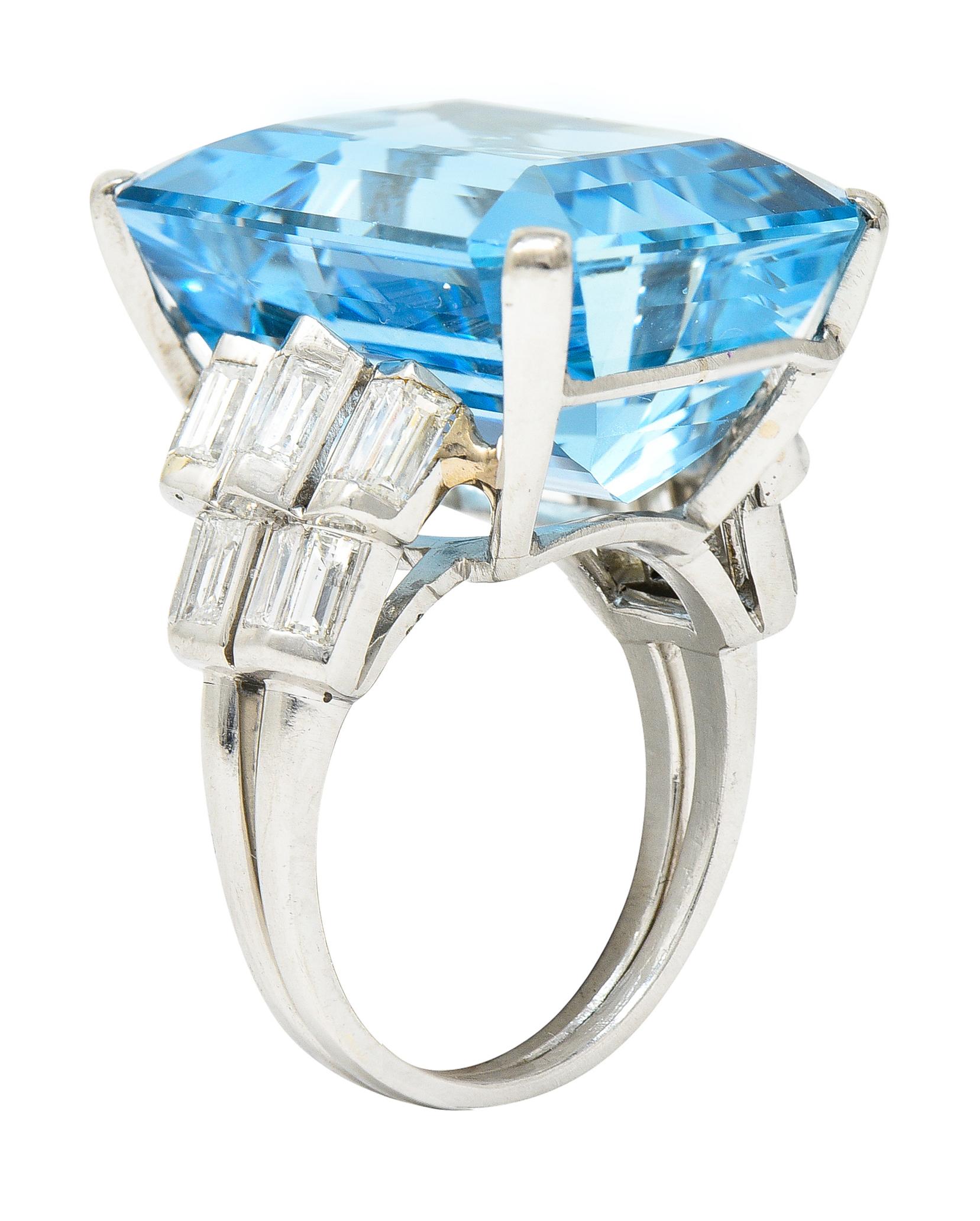 Centering a substantial emerald cut aquamarine weighing approximately 23.76 carats total. Transparent light to medium blue in color - prong set in wire basket. Flanked by stepped chevron motif cathedral shoulders. Bar set throughout with baguette