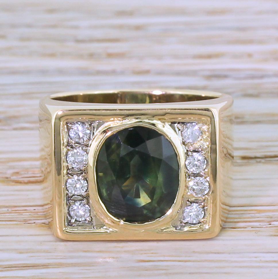 A lovely and bold green sapphire ring. The large, olive coloured centre stone is rubover set in yellow gold and flanked either side by a line of four white and bright round brilliant cut diamonds. The rectangular shaped head of the ring conforms to