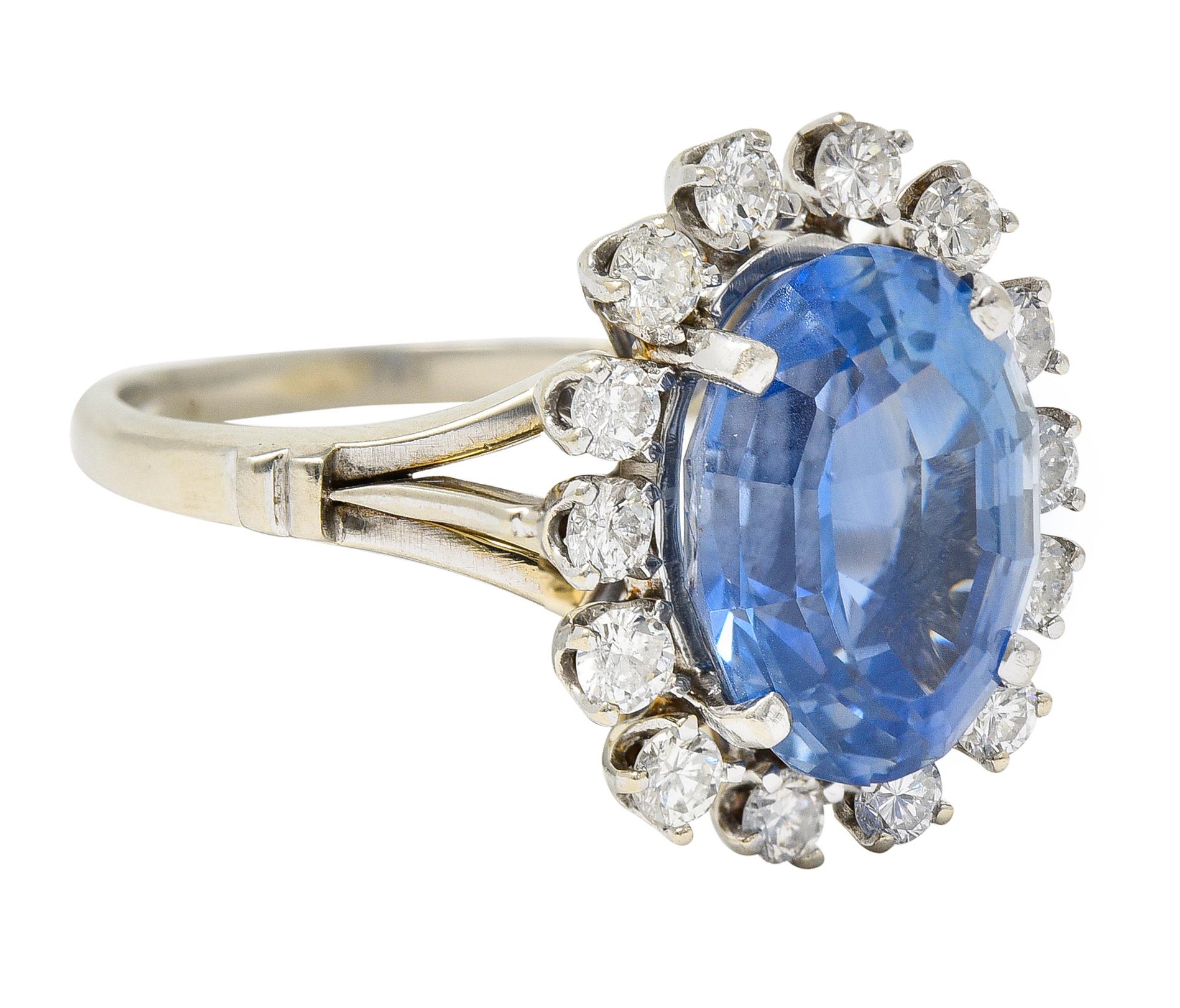 Centering an oval step cut sapphire weighing 6.47 carats - transparent light cornflower blue. Natural Ceylon in origin with no indications of heat treatment - prong set. With a halo surround of round brilliant cut diamonds. Weighing approximately