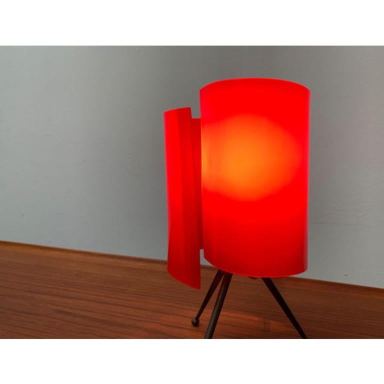Small mid century table lamp with red plastic diffuser.

The most striking detail of this luminaire is the curved umbrella made of translucent red plastic. The umbrella sits on an elegant brass tripod foot. The feet conclude spherical at the bottom