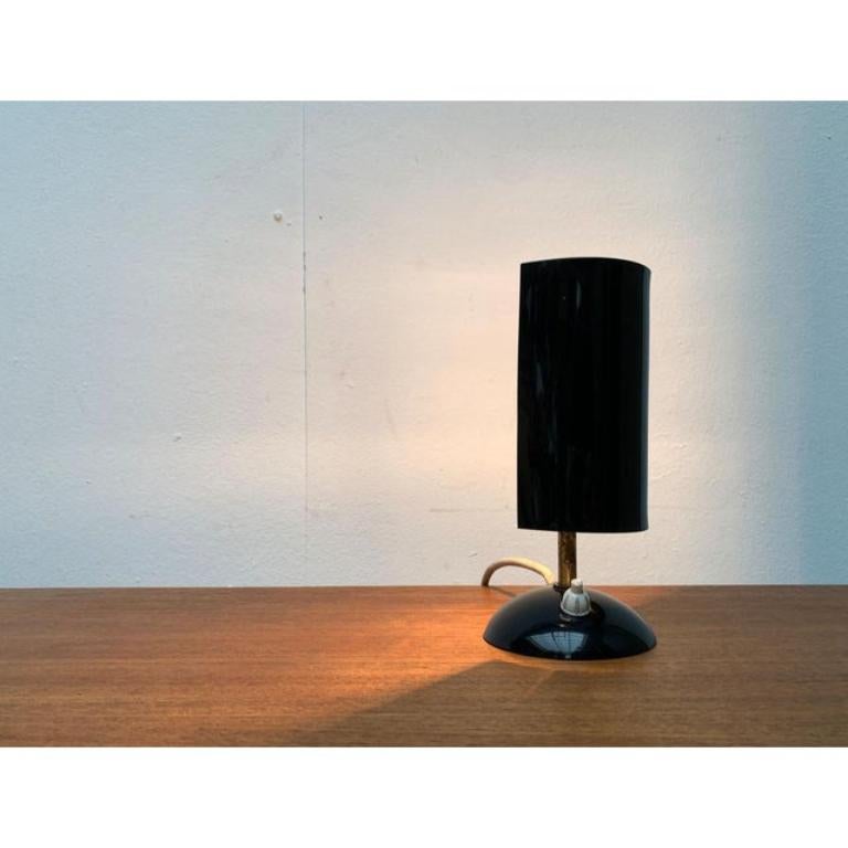 Petite lampe de table avec réflecteur en acrylique.

Le détail le plus frappant de cette lampe est l'abat-jour en forme de tonneau en acrylique noir. Celui-ci entoure la douille de la lampe et peut tourner autour de l'illuminant. Cela permet