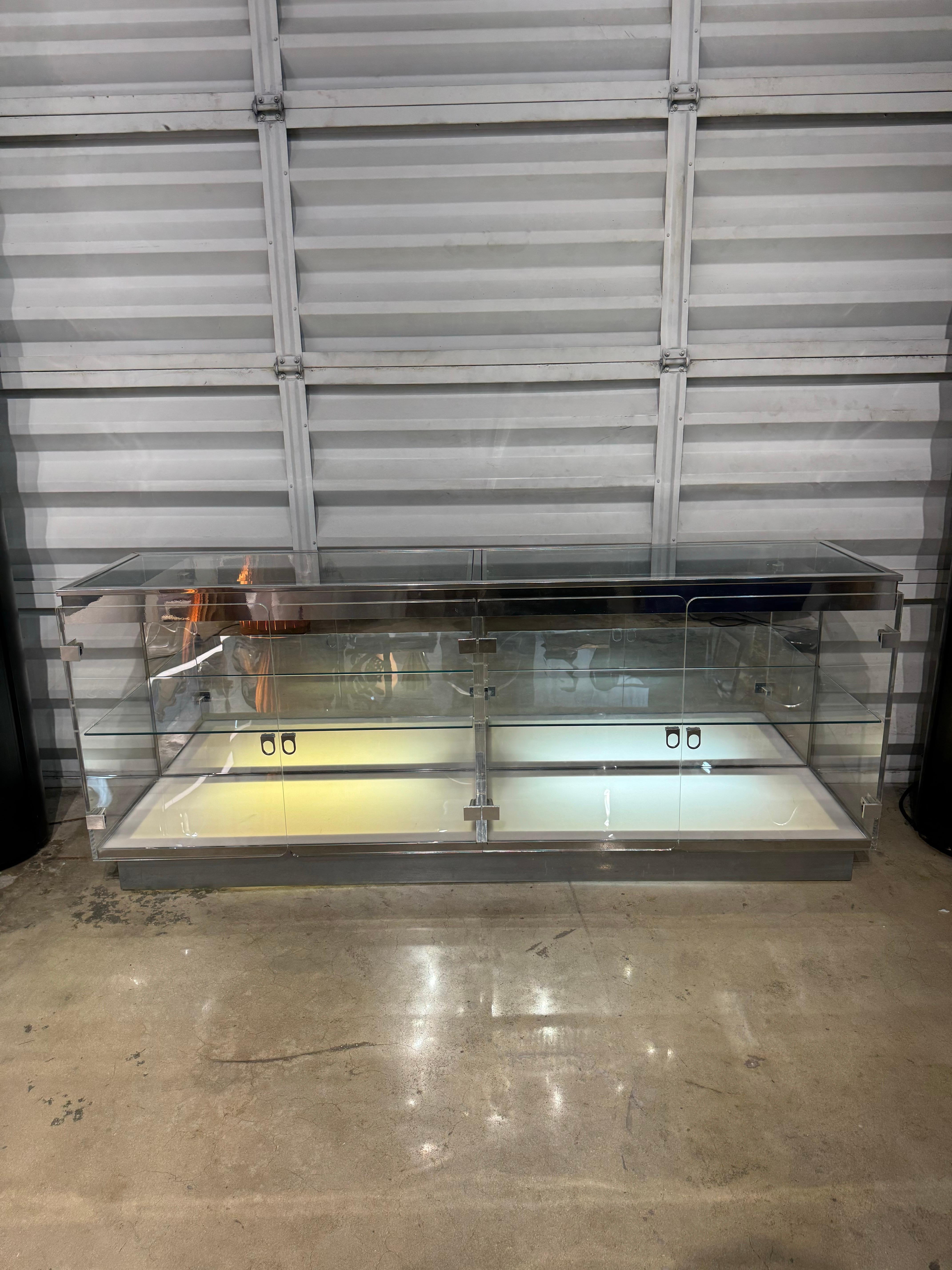 1970s mid-century modern lucite sideboard with glass interior shelves, glass top and transparent plexiglass outer shell with chrome accents and plexi doors.  The display sideboard also has fluorescent lamps under an opaque acrylic sheet to
