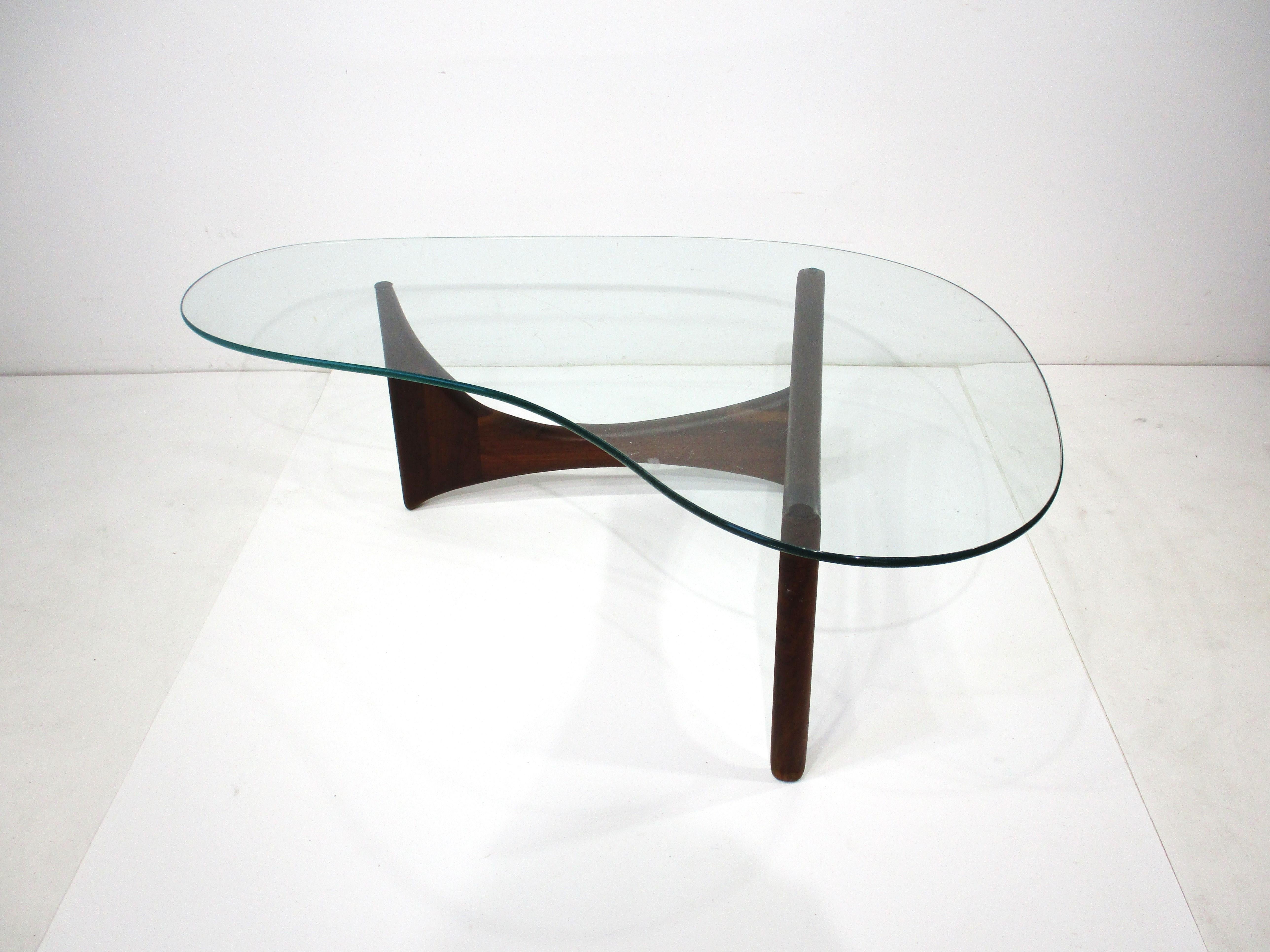 A solid dark walnut based coffee table with biomorphic cut glass top having rounded edges . The base folds and adjusts to the way you want the bottom to look in your interior designed by Adrian Pearsall for his company Craft Associates . 