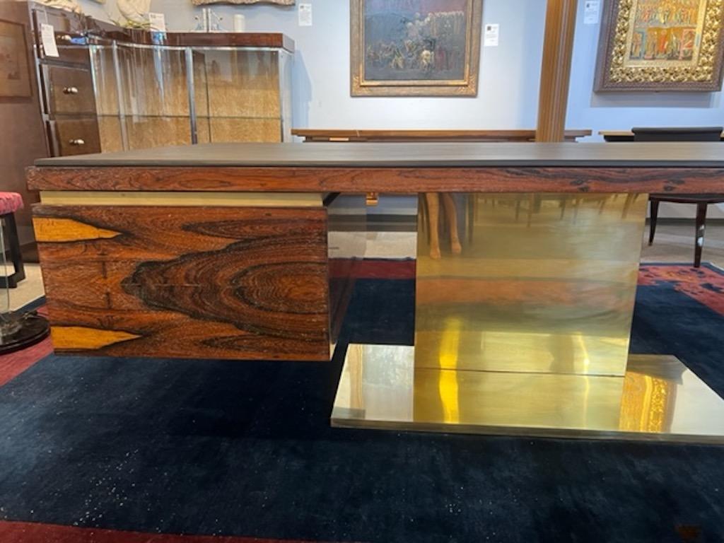 Desk is made out of palisander wood. Table top is reupholstered with brown cowhide. There is wide drawer for files on the left side and smaller drawer on the right. Desk is elevated by wide and prominent brass base. 
Desk done by famous American