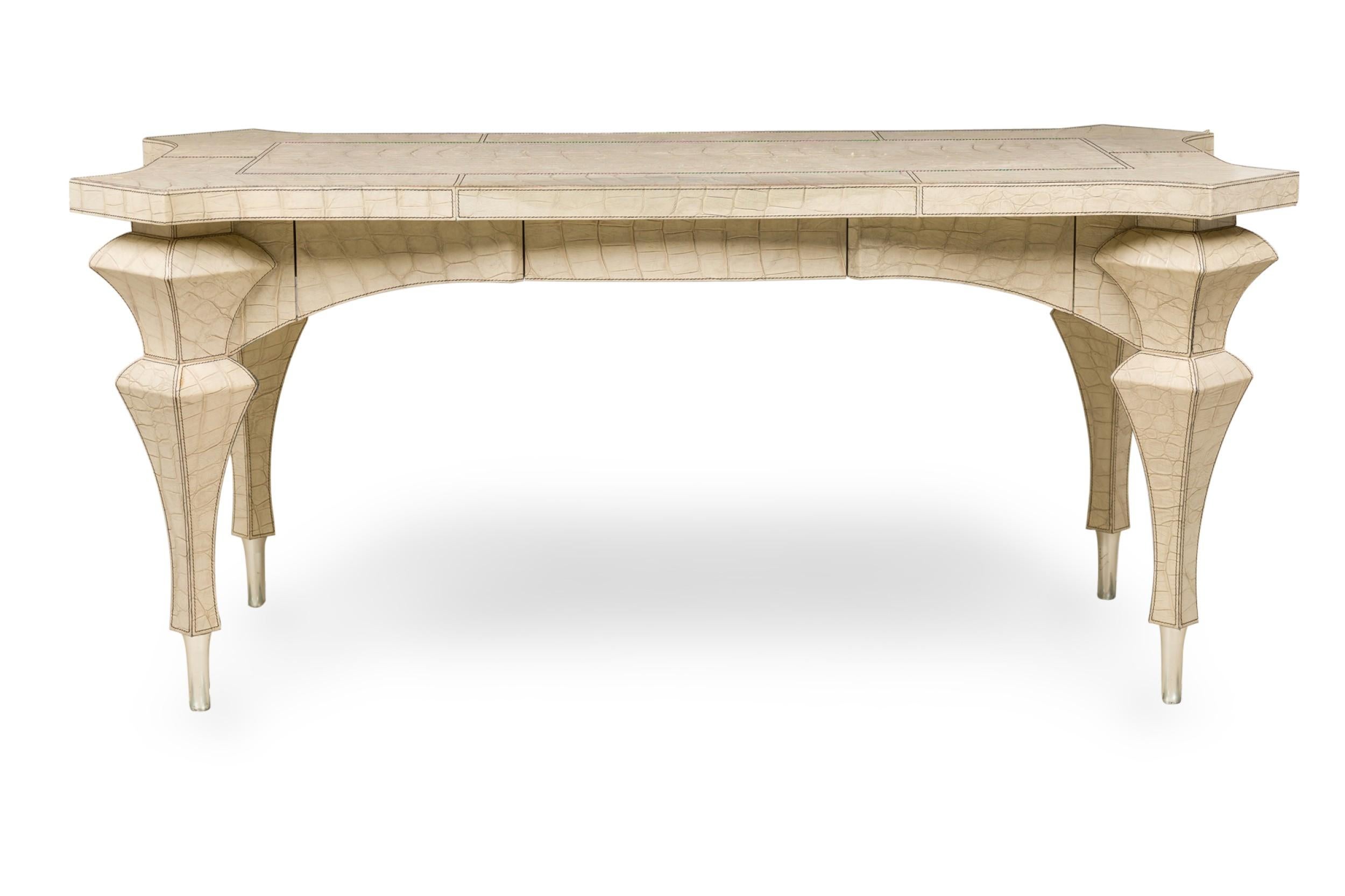Mid-Century American table desk having an embossed off-white faux reptile leather veneer featuring a shaped top with canted corners and matching sculptural legs resting on 4 exposed silver metal tapered legs. (KARL SPRINGER) (Companion pieces: