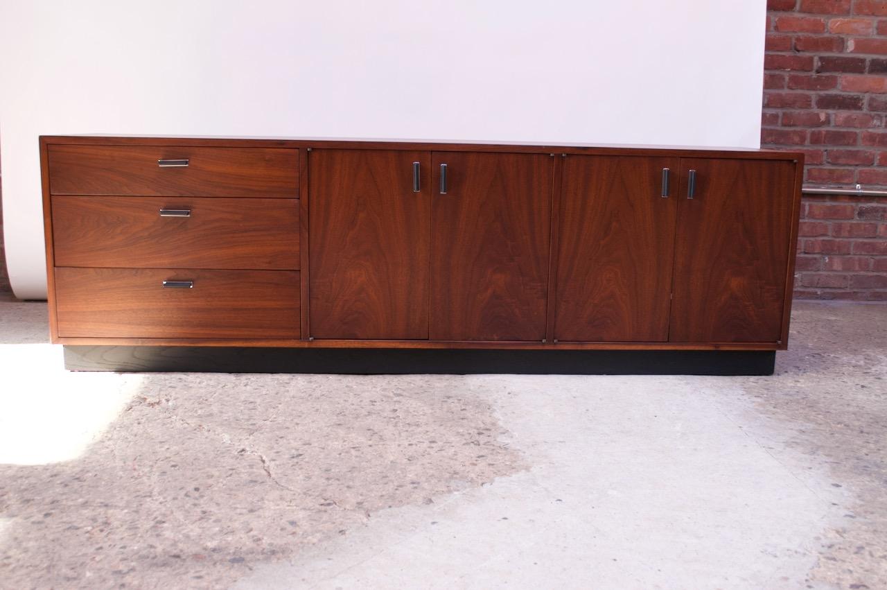 Midcentury American credenza composed of a walnut frame with an ebonized plinth-base and ebonized aluminum pulls with chrome-plated platforms.
Features three drawers to the left and two cabinets to the right, each interior space flanked by an