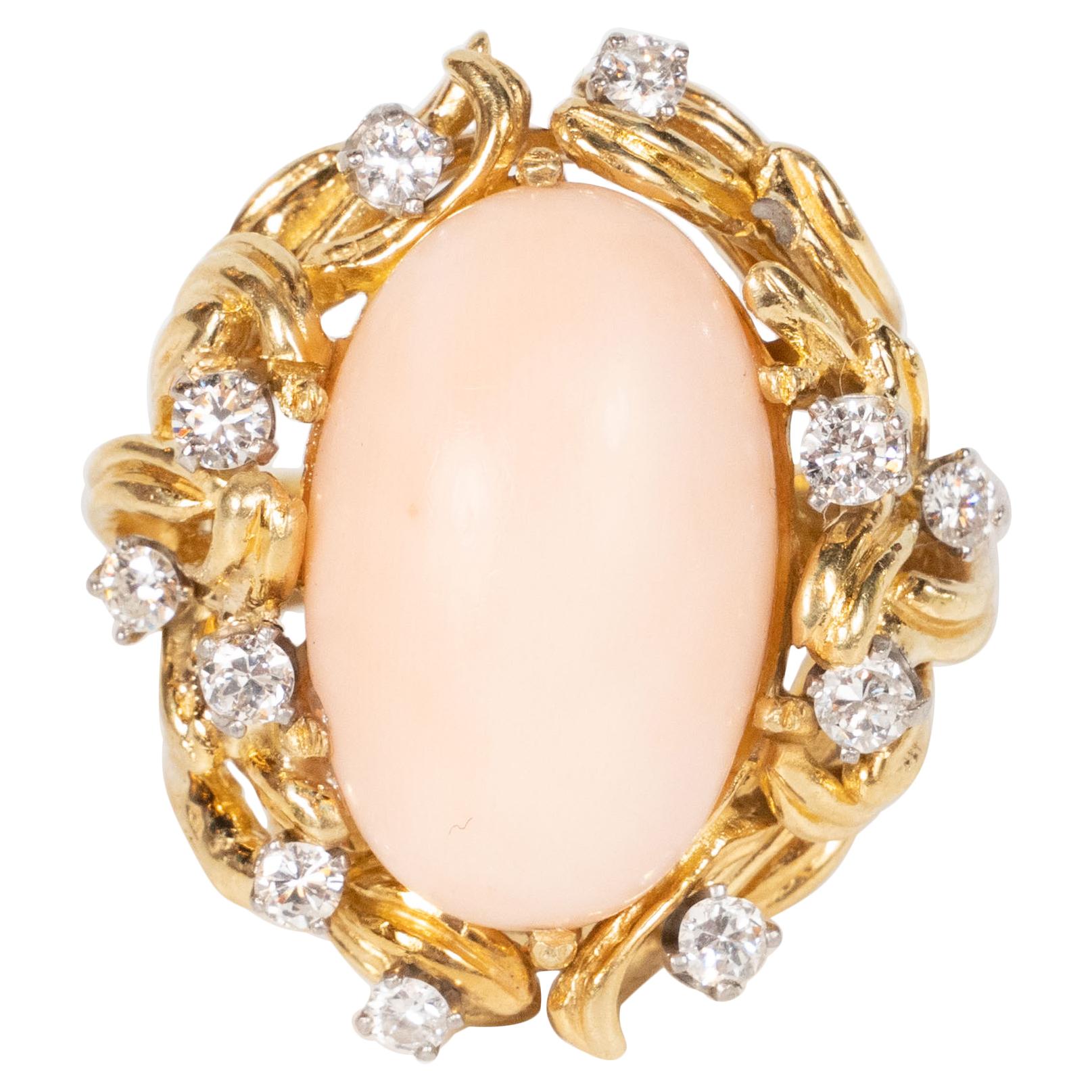 This Mid-century cocktail ring was hand crafted in the United States, circa 1960. features a domed angel coral stone set in a 14k yellow gold designed with stylized foliate tendril details supporting 12 full cut fine diamonds. This ring is