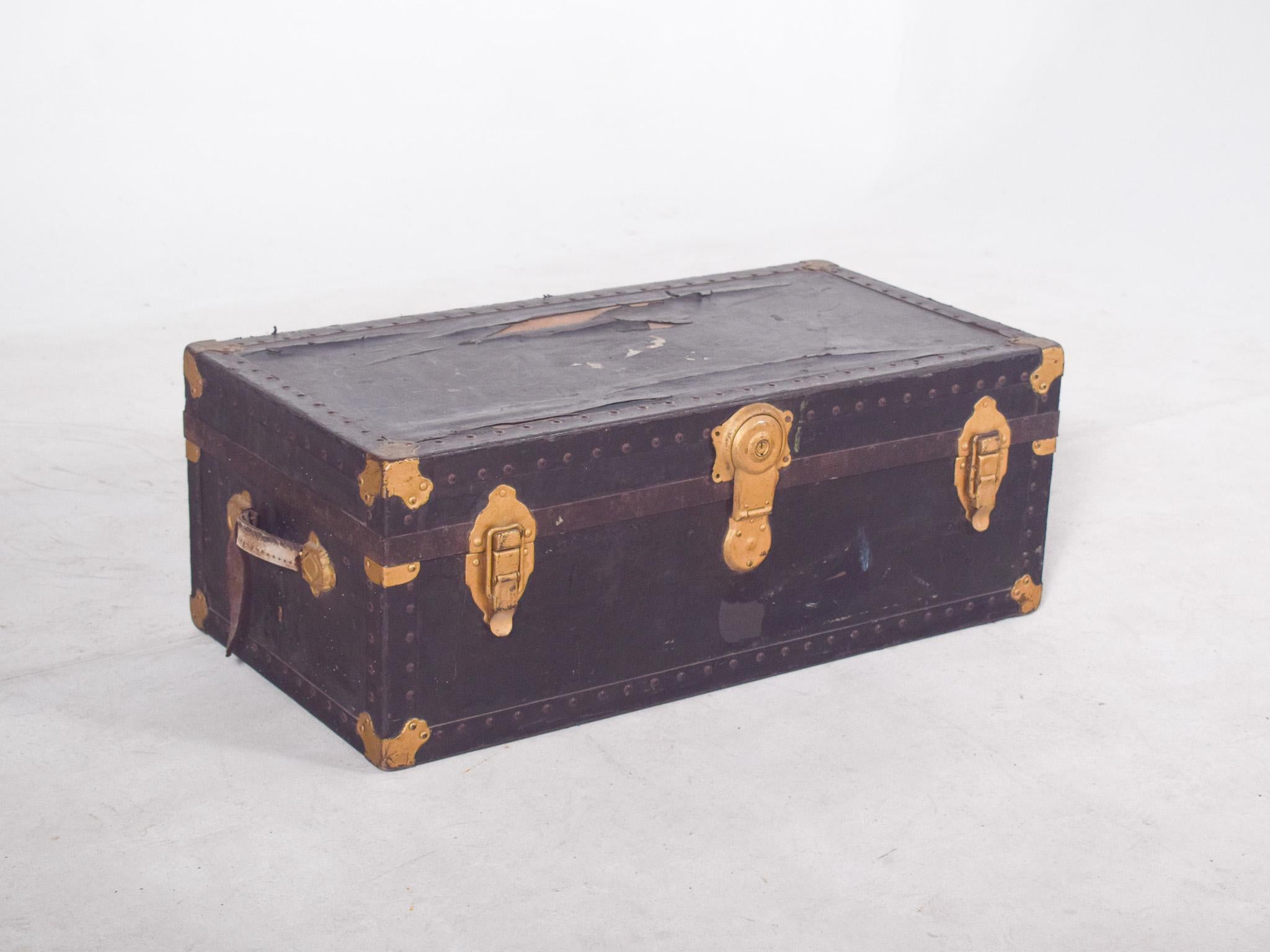Bolsa de viaje antigua forrada de cuero negro. Con herrajes metálicos de latón dorado en los bordes, esquinas y cremalleras. La estructura está reforzada con tacos en todo su perímetro, lo que le confiere robustez y carácter decorativo. Tiene un asa
