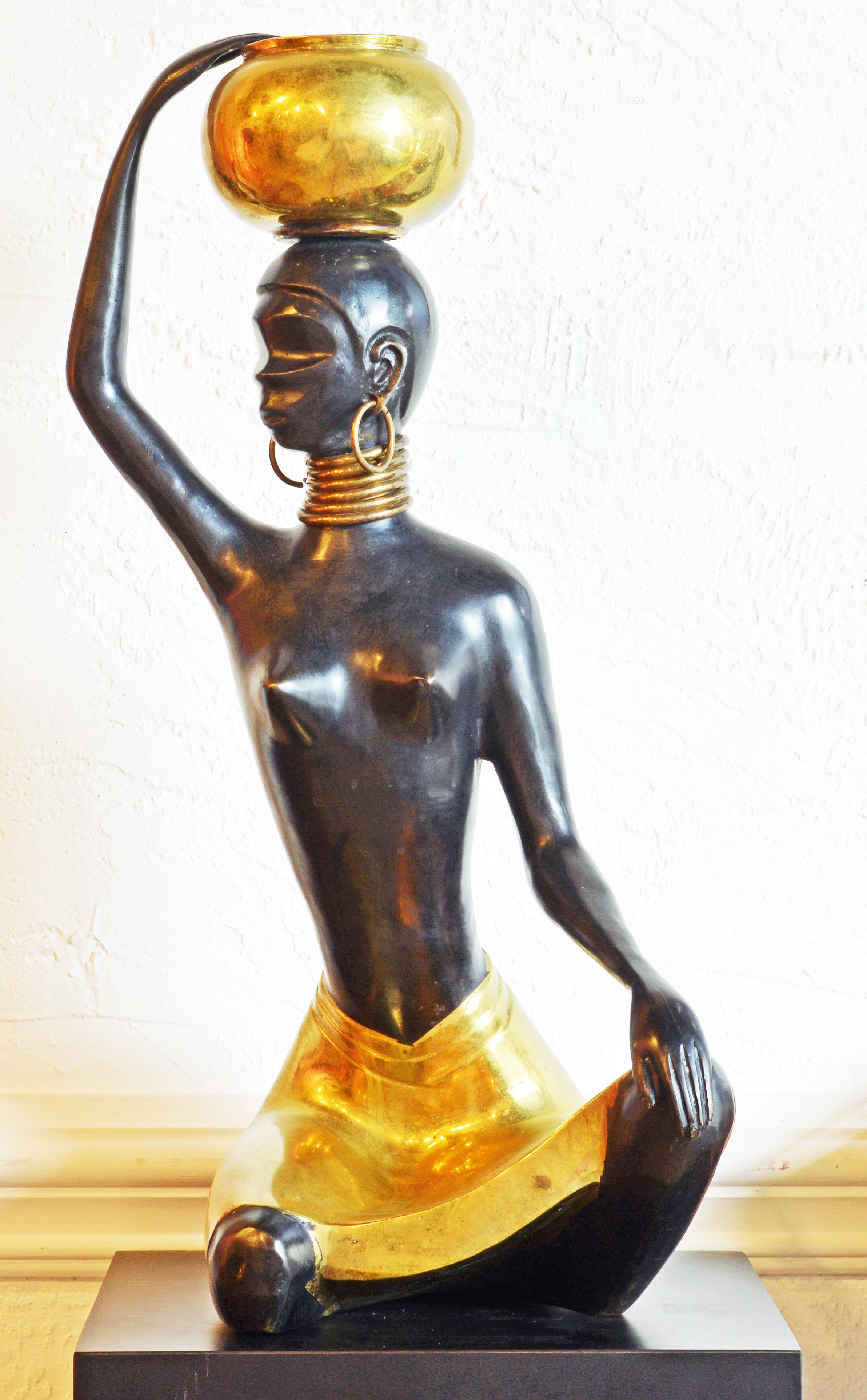 This Art Deco style patinated bronze and brass sculpture reminiscent of Hagenauer's style features a young African woman sitting in a brass skirt legs crossed with her right hand balancing a brass vessel on top of her head. The face, hands and