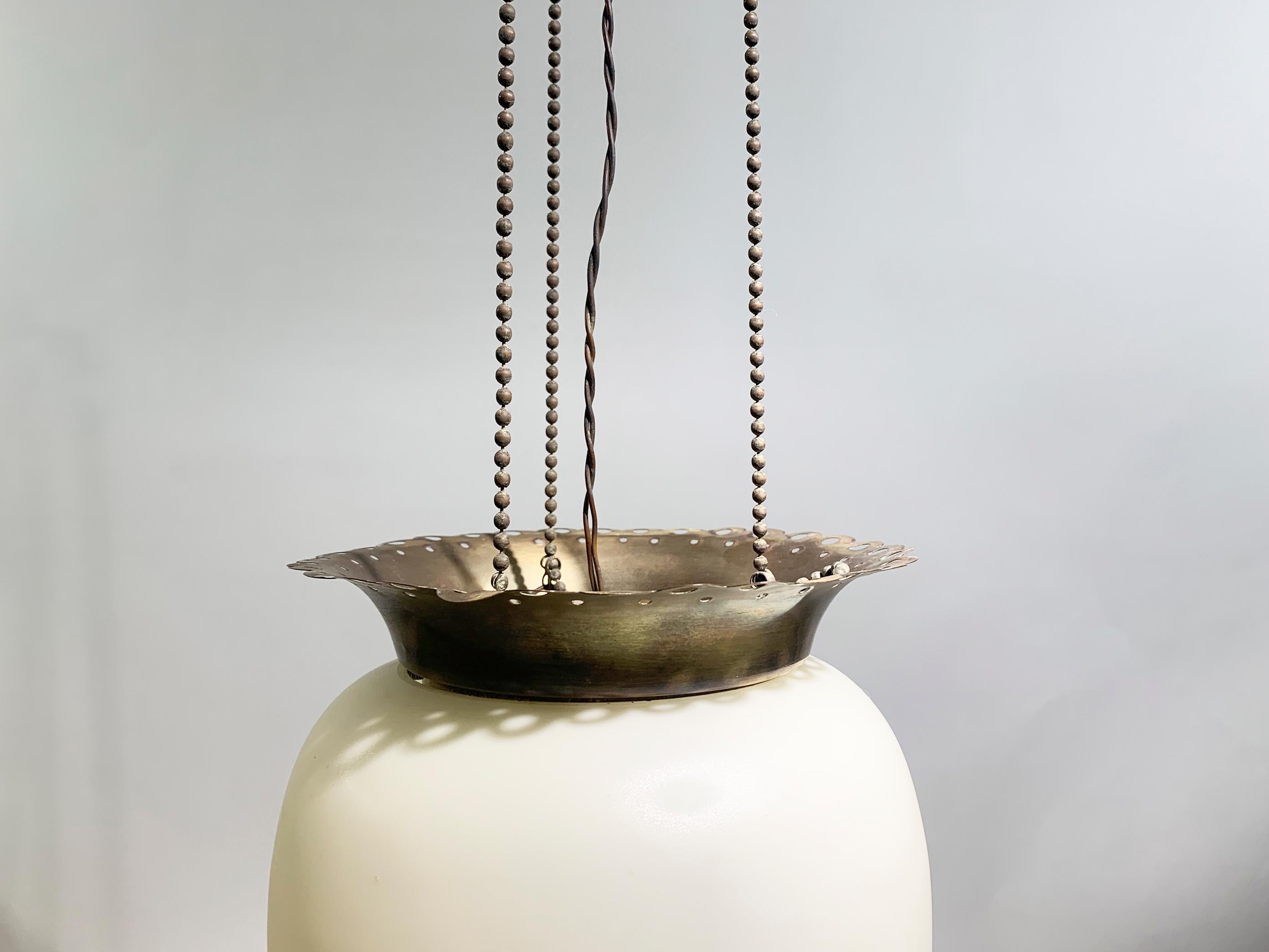 This pendant features an elongated, egg-shaped opalized glass, which gives it a soft, diffused glow when lit. The shade is held in place by ornate chiseled bronze fittings at both the top and bottom, with scalloped or filigree-like edges, adding a