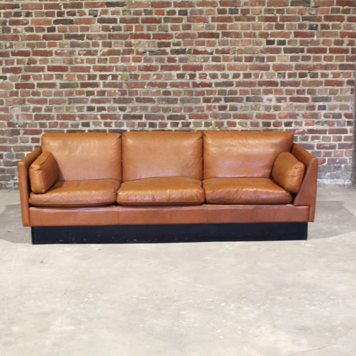 Very original asymmetrical sofa in brown leather. This beautifully patinated sofa rests on a blackened wooden base. The leather is thick and supple. Of Scandinavian origin, it has the interesting feature of being slightly asymmetrical in the