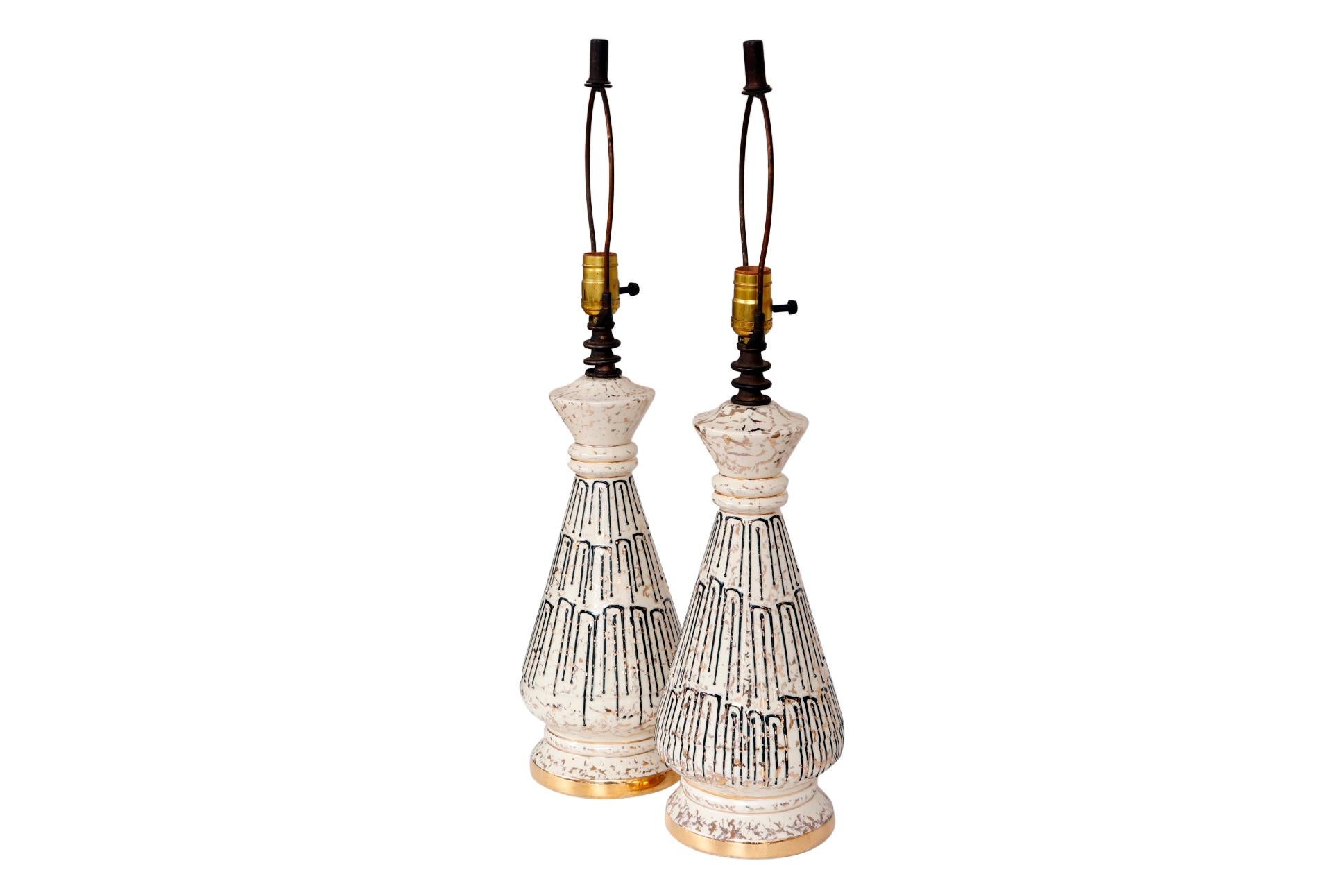 A pair of mid century atomic style ceramic lamps in white, black and gold. Round lamps that taper upwards are decorated with rectangular dripped black lines, mirrored on the shades, and flecked with gold. Bases are trimmed with gold bands. Each lamp
