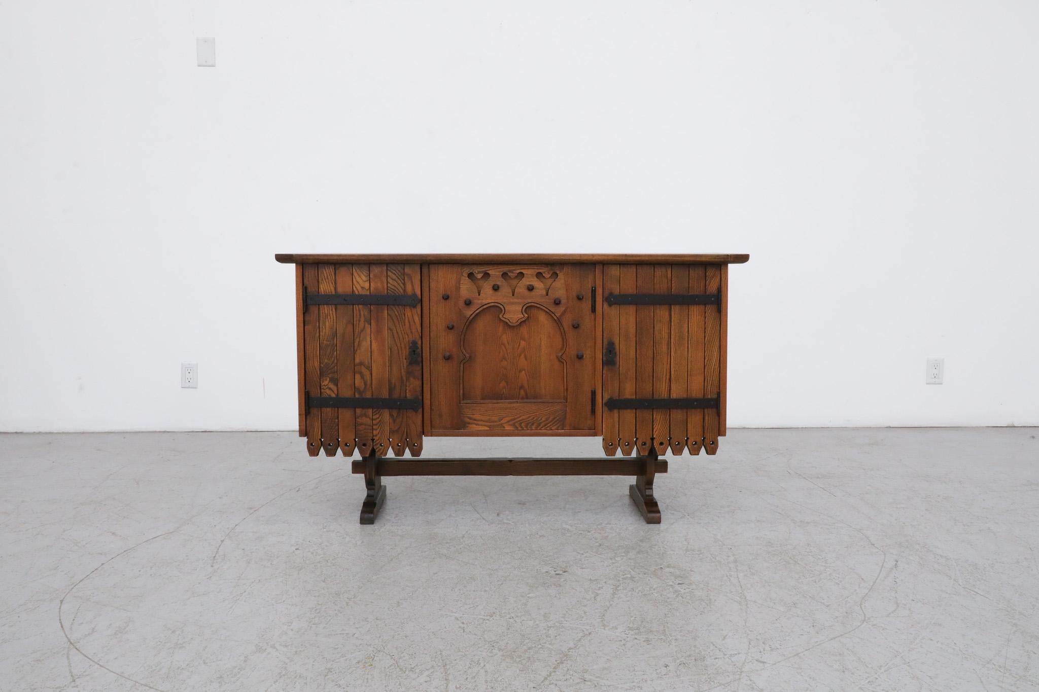 Mid-Century brutalist wood credenza with dark stained tone, ornate detail and iron hardware. This cabinet has interior shelving and a little bar area behind the door with the three heart cut-outs on the door. Both the left and the right door have
