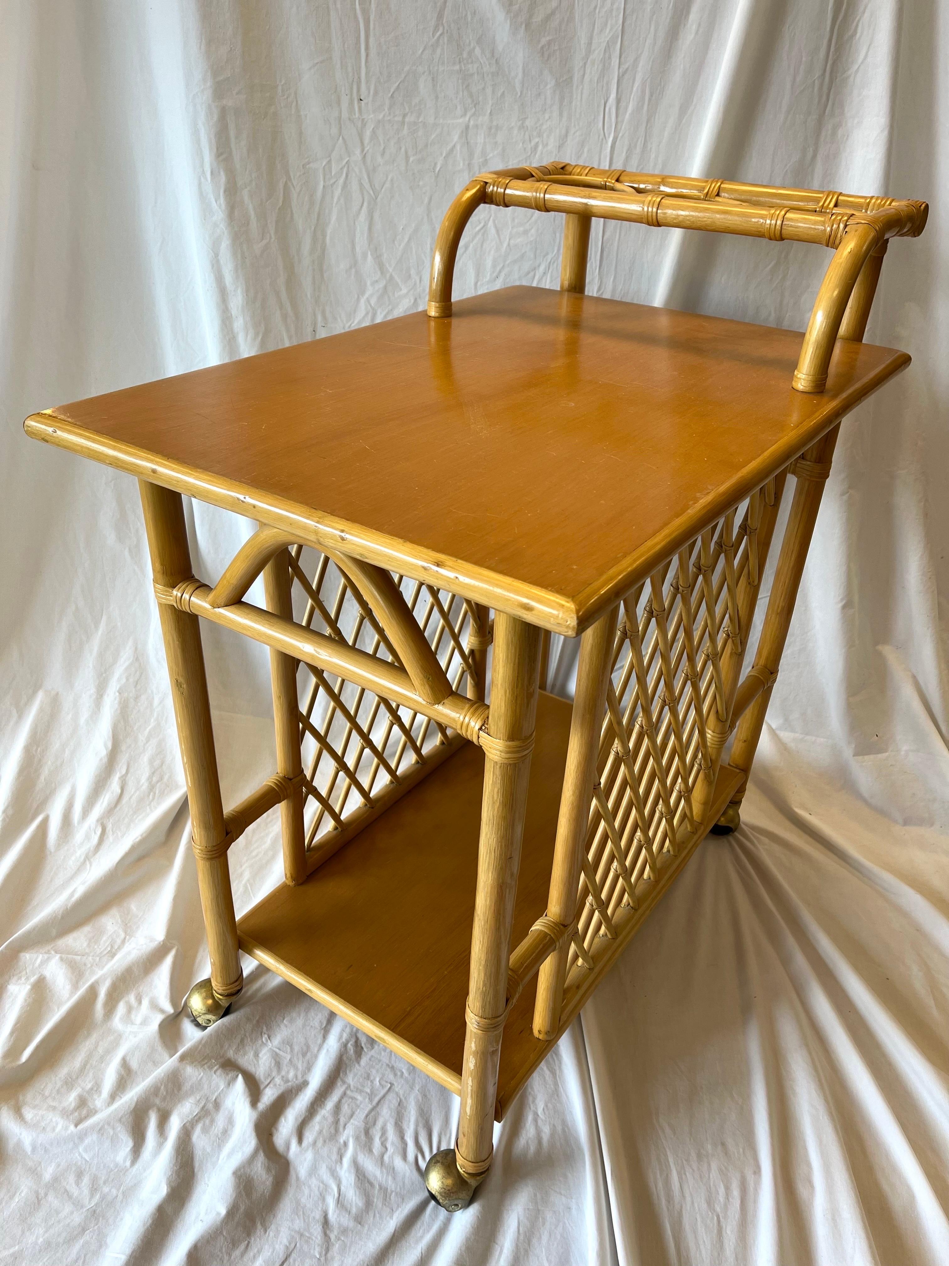 A vintage, mid 20th century bamboo, two tier bar cart or trolley with a cross hatched or trellis design on either side as well as a handle that incorporates storage for three bottle os your favorite beverage. The top shelf (no pun intended) is well
