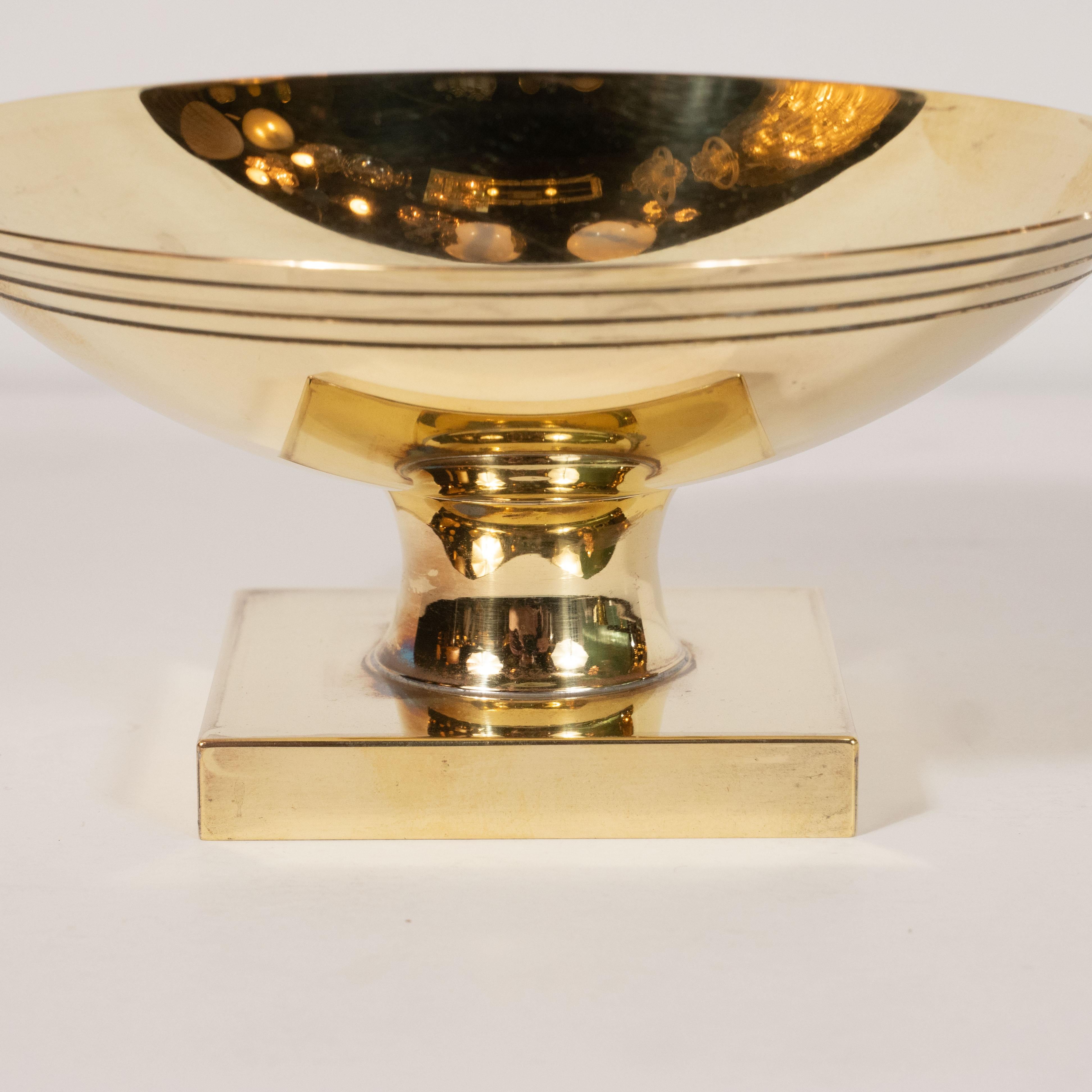 This stunning Mid-Century Modern dish was realized by the esteemed Tommi Parzinger for Dorlyn Silversmiths, circa 1950. It features a concave circular top with three bands circumscribing the perimeter. The piece sits on a volumetric square base that